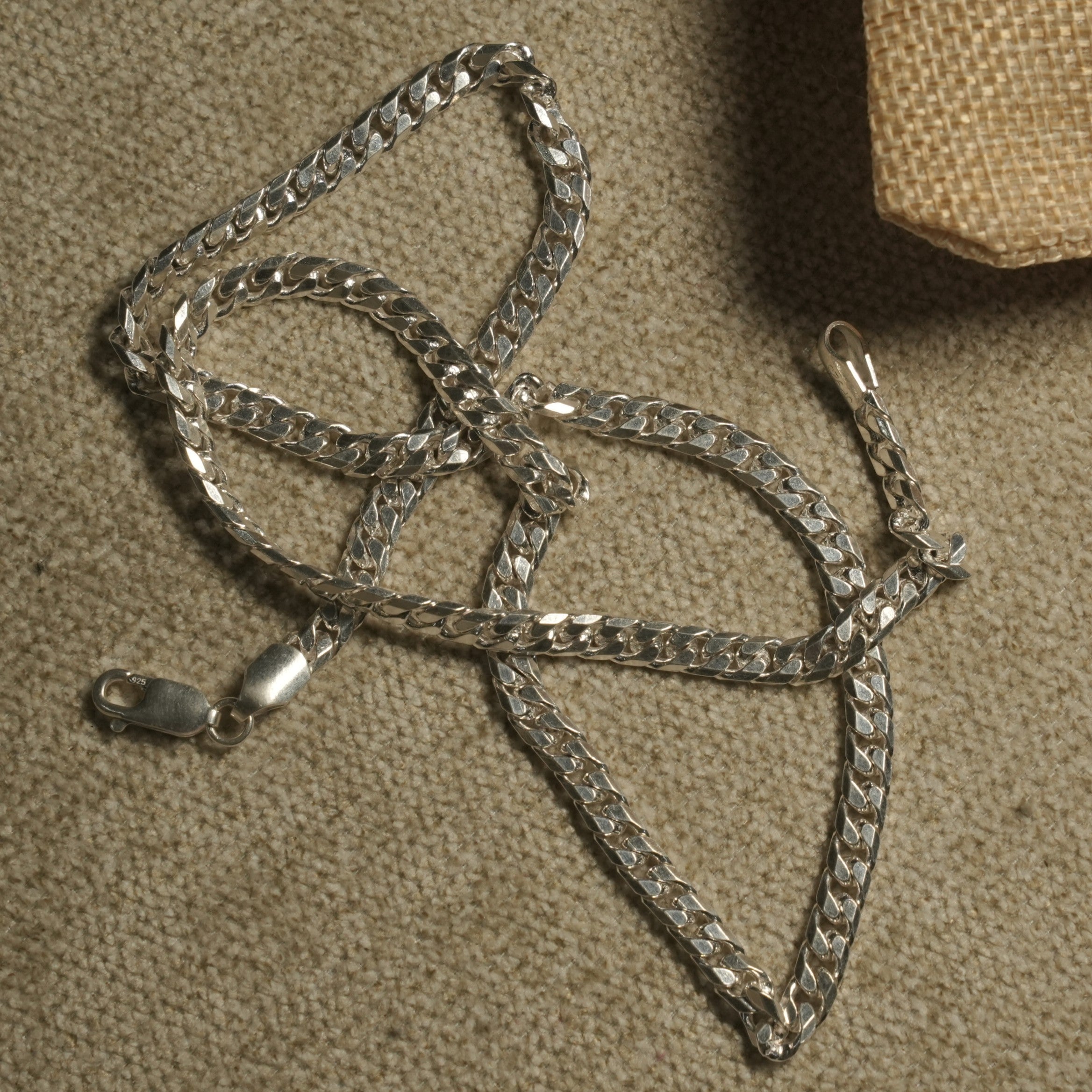 Bevelled sterling silver curb chain with flat interlocking links arranged on beige fabric beside a woven jute pouch, lobster clasp open.