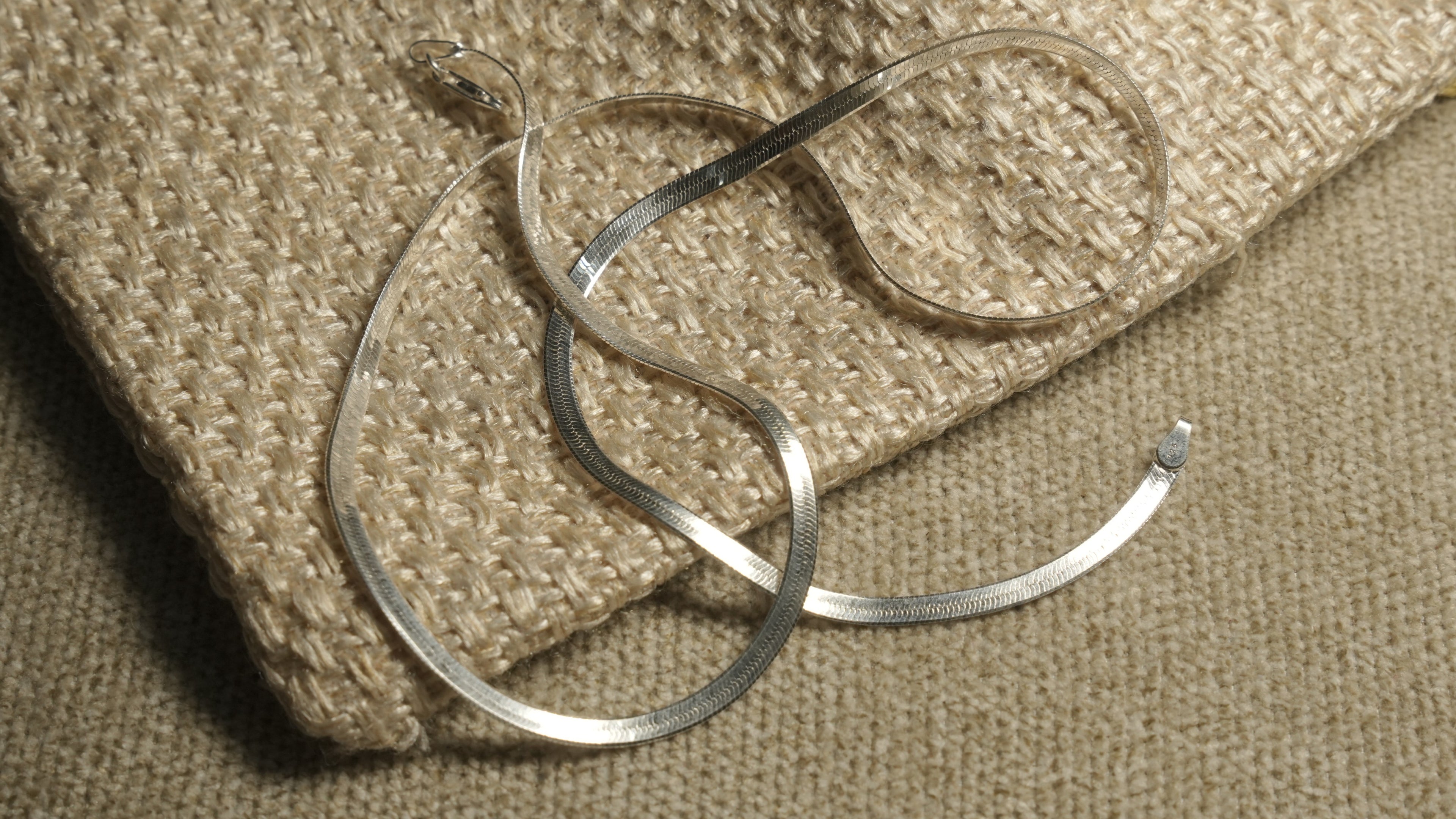 Flat sterling silver herringbone chain draped in soft curves across the corner of a woven jute pouch.