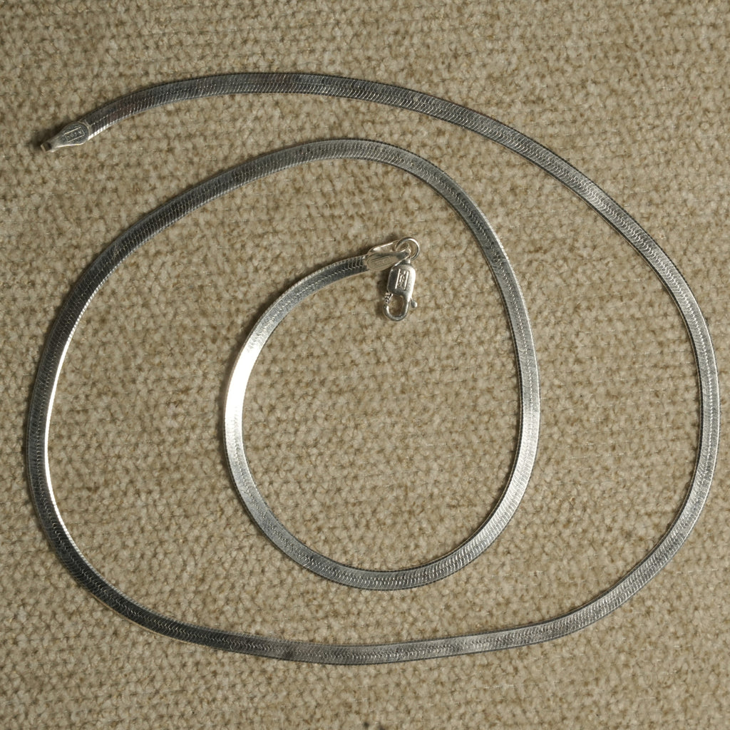 Herringbone chain coiled in a loose spiral on beige fabric, its lobster clasp resting at the centre.