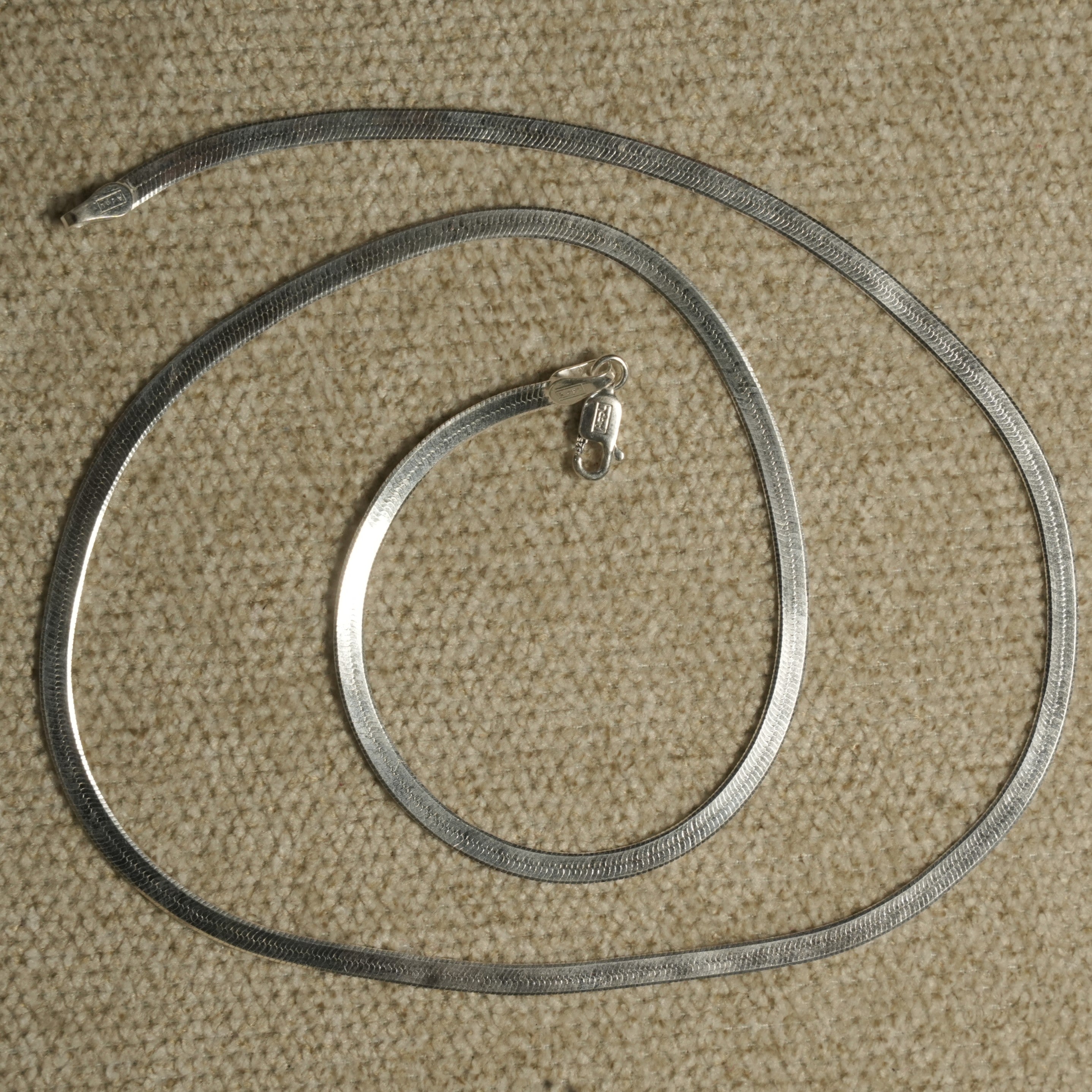 Herringbone chain coiled in a loose spiral on beige fabric, its lobster clasp resting at the centre.