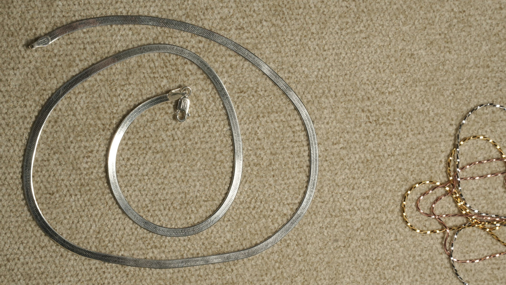 Sterling silver herringbone chain coiled on a beige textile, with fine tri-tone chains visible at the edge of the frame.