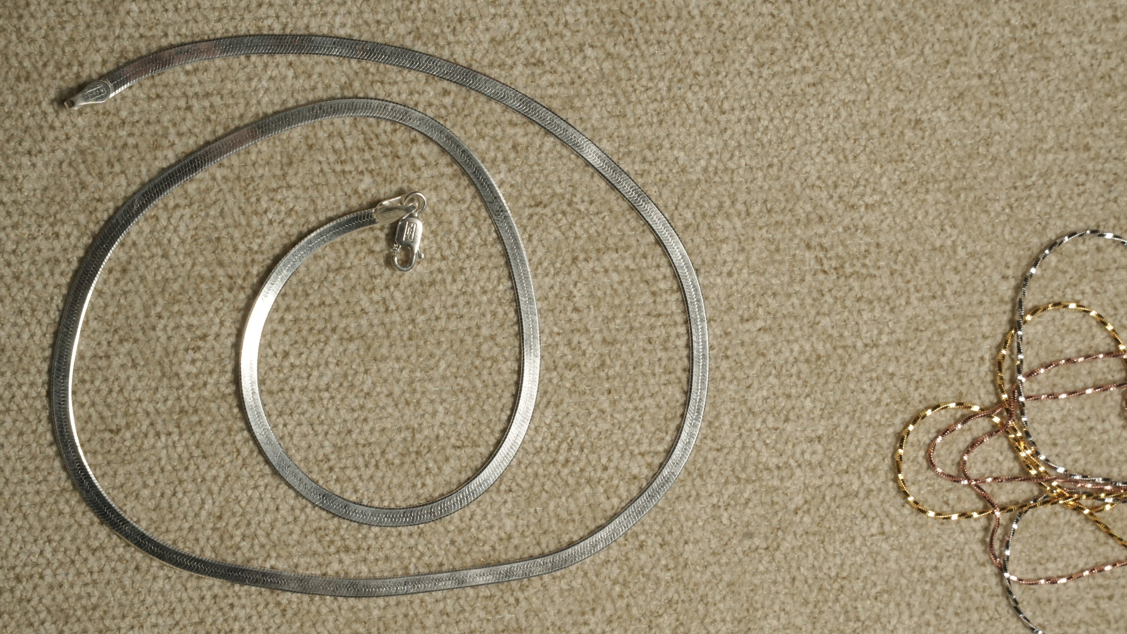 Sterling silver herringbone chain coiled on a beige textile, with fine tri-tone chains visible at the edge of the frame.