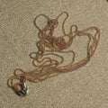 Multiple fine ball-bead chains in silver, gold-tone and rose gold-tone gathered on a beige textile, with a woven jute pouch and gold-tone cord above.