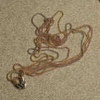 Multiple fine ball-bead chains in silver, gold-tone and rose gold-tone gathered on a beige textile, with a woven jute pouch and gold-tone cord above.