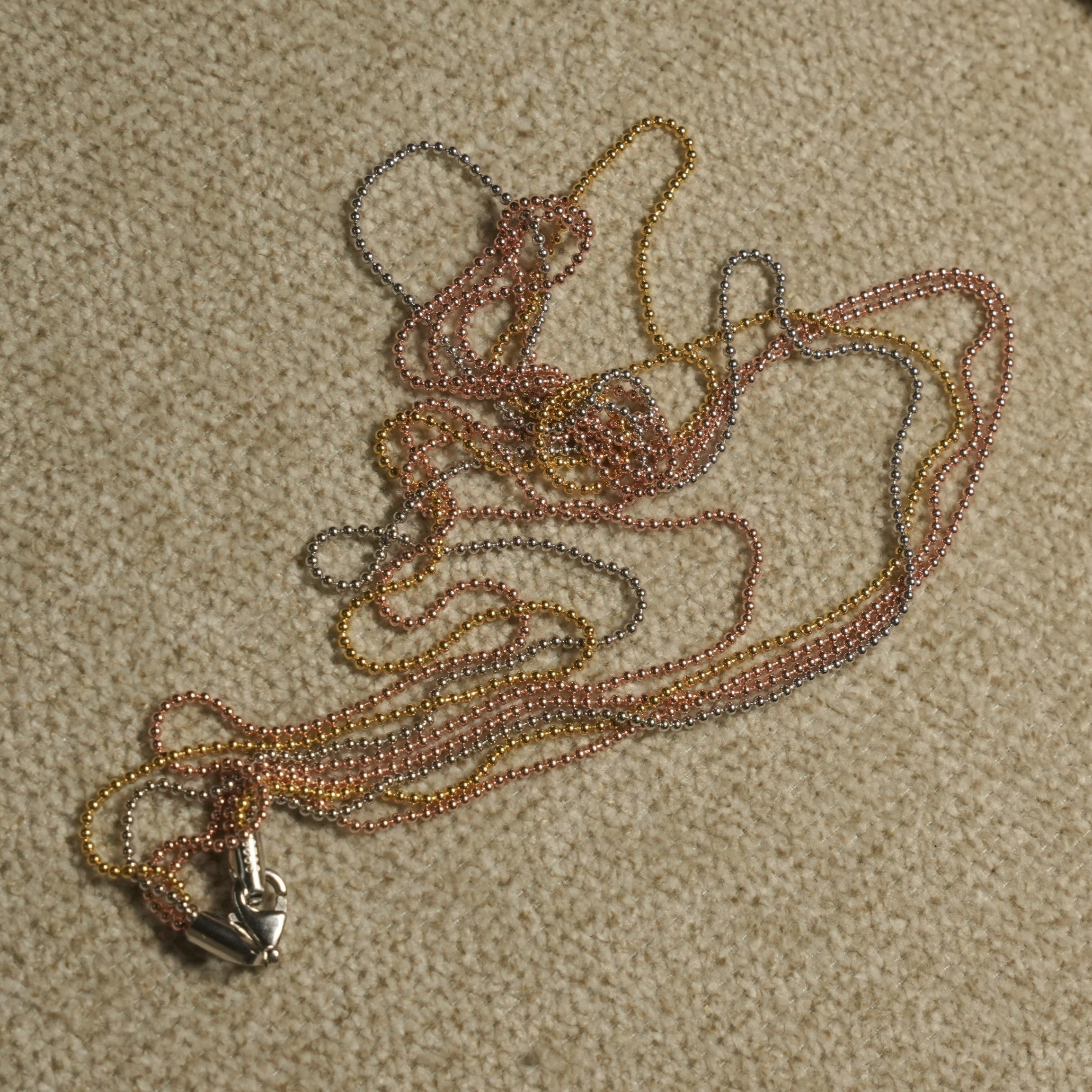 Multiple fine ball-bead chains in silver, gold-tone and rose gold-tone gathered on a beige textile, with a woven jute pouch and gold-tone cord above.