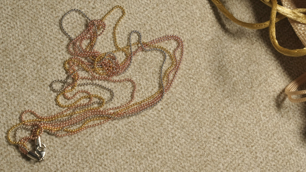 Tri-tone multi-strand bead chain resting in soft loops on beige fabric, its lobster clasp at the lower left and a jute pouch at the edge.