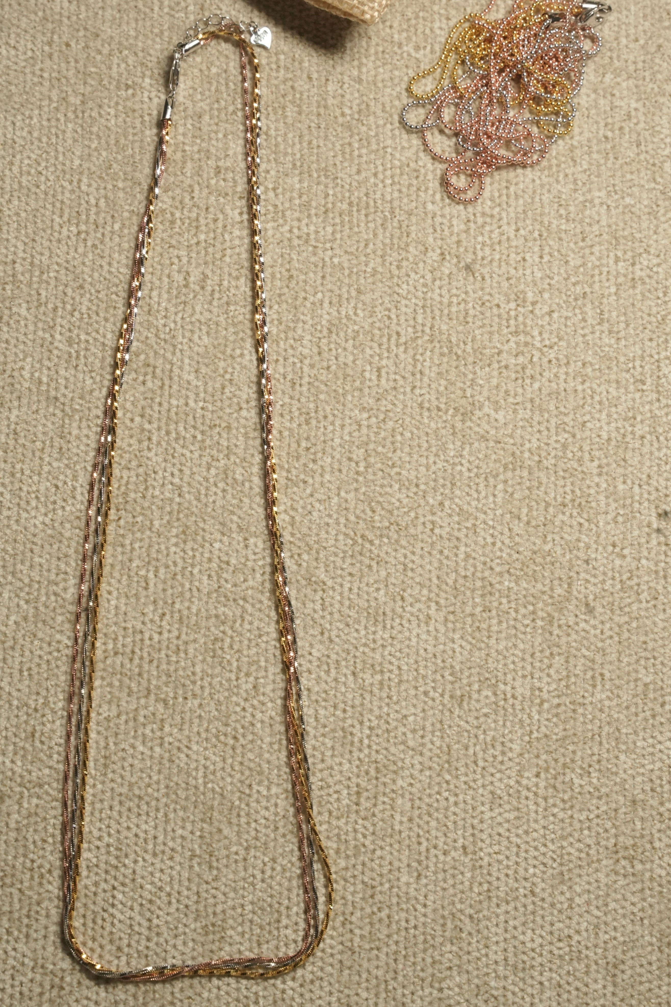 Three long twisted serpentine chains in silver, gold-tone and rose gold-tone laid in a narrow loop on beige fabric, clasp and extender at the centre.