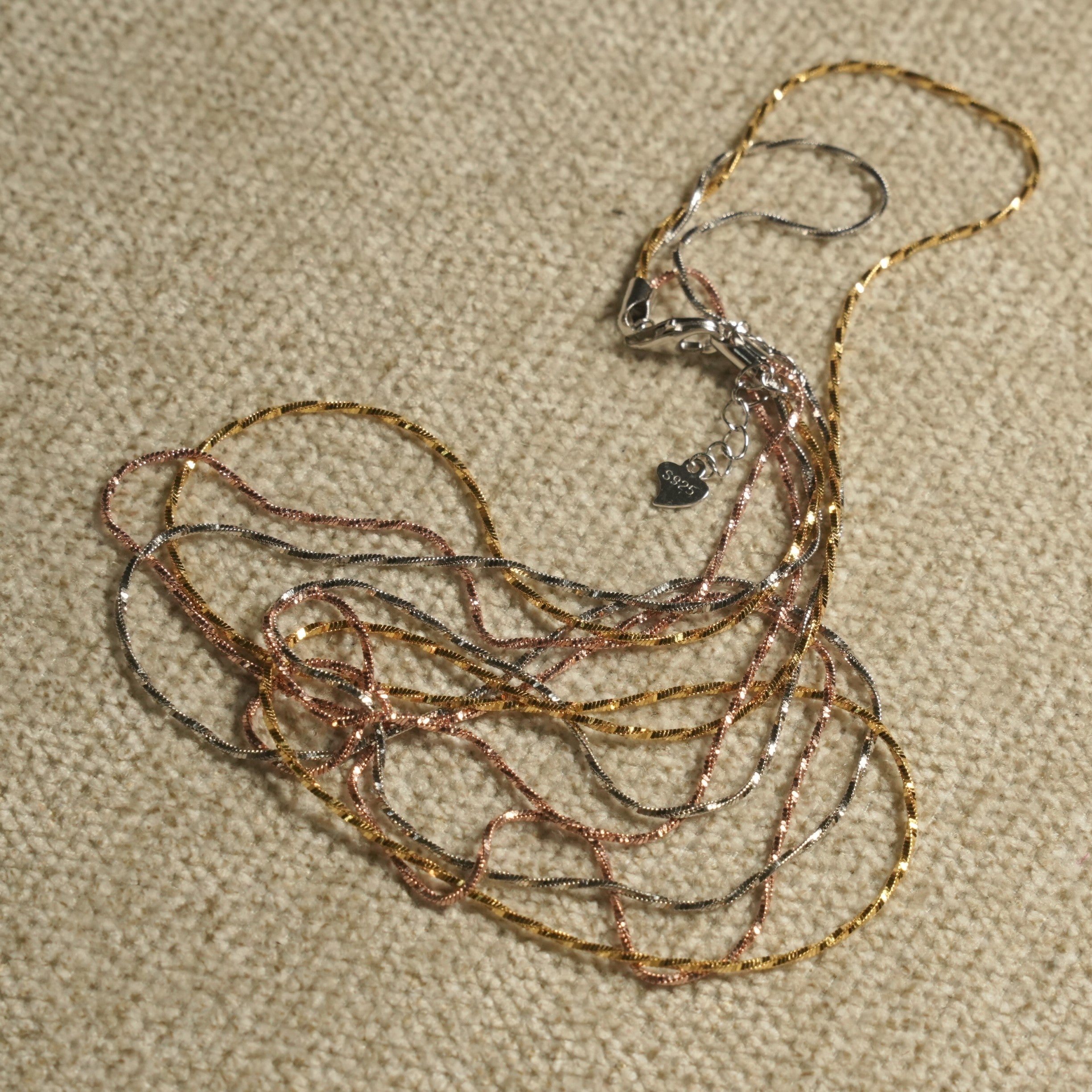 Tri-tone twist chain gathered in soft loops on a beige textile, showing the lobster clasp, extender chain and heart tag stamped 925.