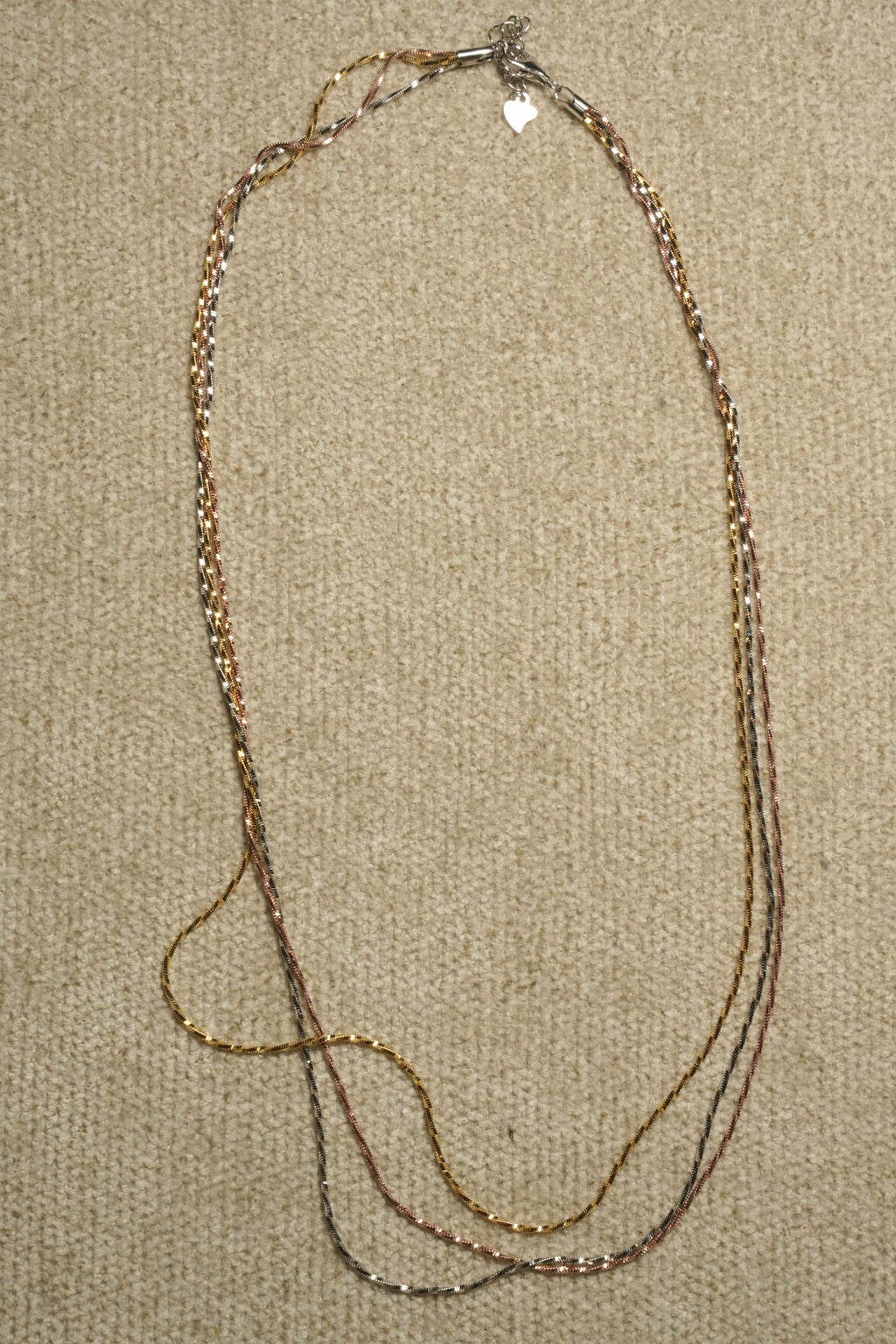 Three long twisted serpentine chains in silver, gold-tone and rose gold-tone laid in a narrow loop on beige fabric, clasp and extender at the centre.