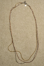 Three long twisted serpentine chains in silver, gold-tone and rose gold-tone laid in a narrow loop on beige fabric, clasp and extender at the centre.