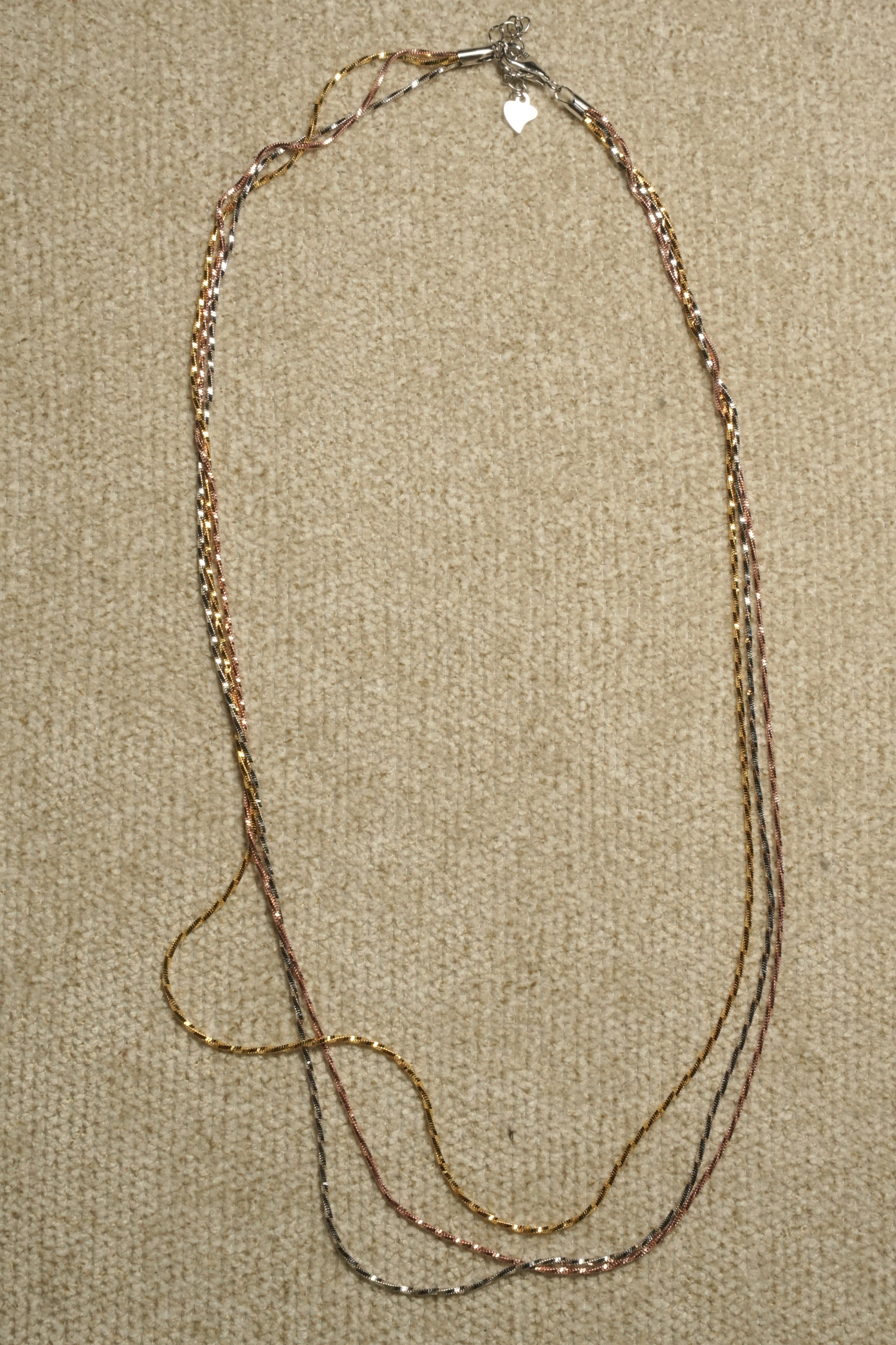 Three long twisted serpentine chains in silver, gold-tone and rose gold-tone laid in a narrow loop on beige fabric, clasp and extender at the centre.