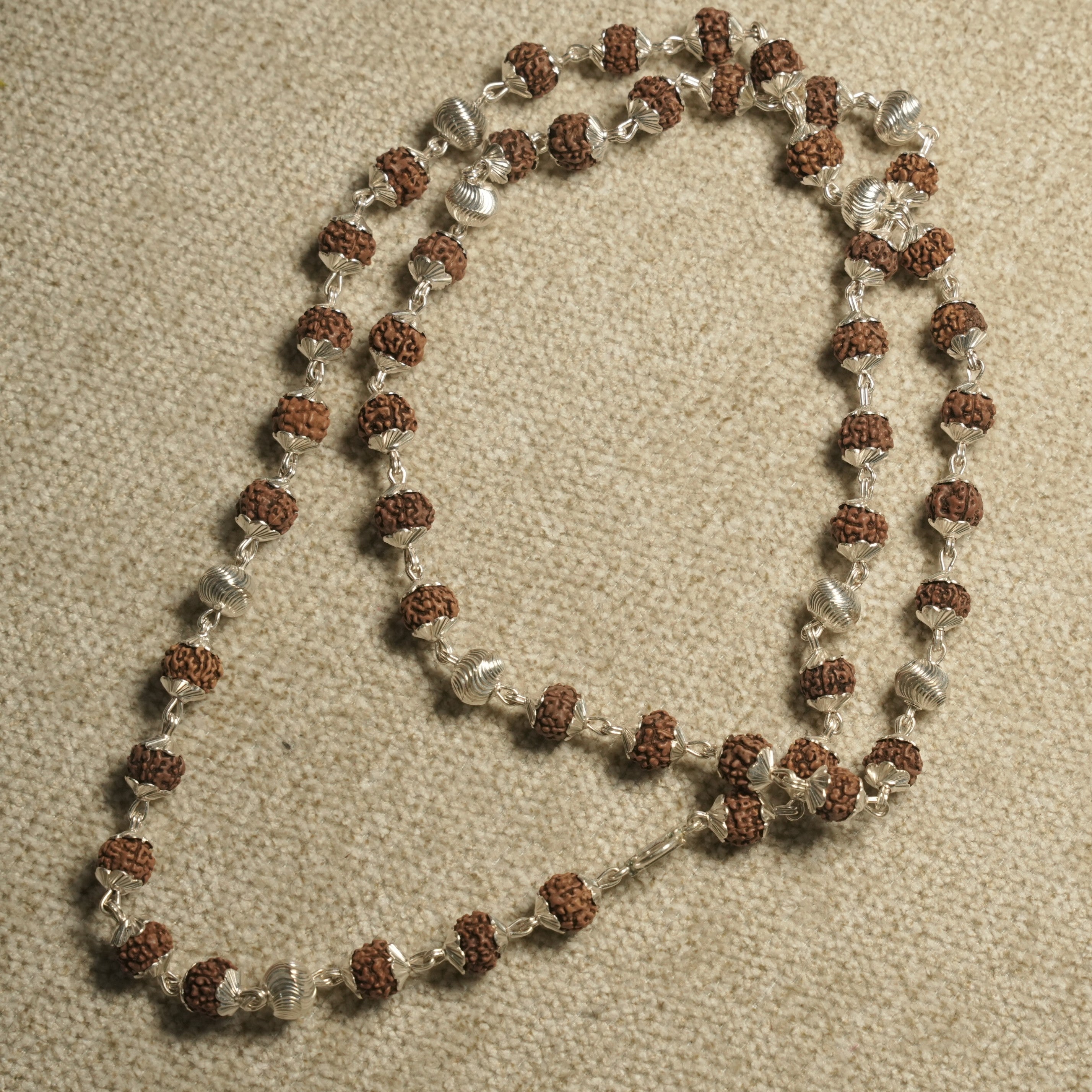 Rudraksha and sterling silver bead chain coiled in a loop on pale carpet with silver ribbed accent beads.