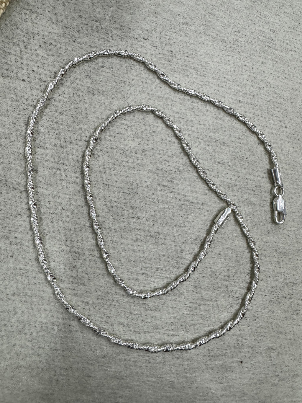 Full 50 cm length of the sterling silver Singapore chain necklace laid in loops on grey felt, with the lobster clasp at the right.