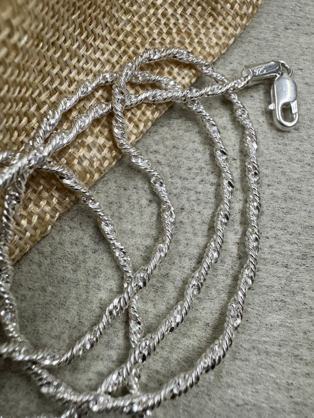 Sterling silver Singapore chain necklace gathered beside a woven jute pouch, the lobster clasp and 925 stamp at the top of the frame.