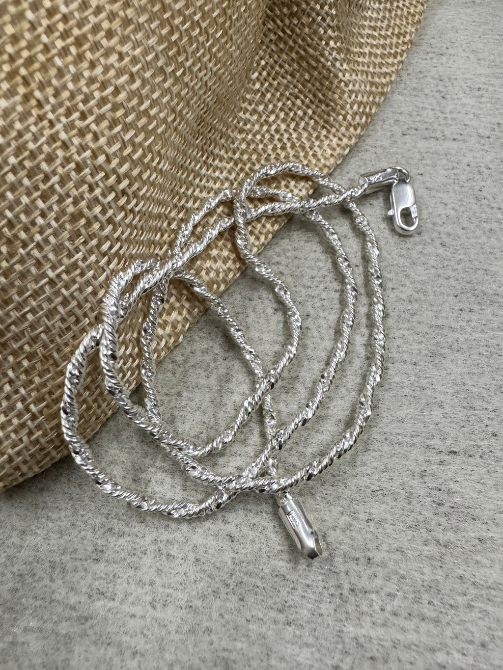 Twisted sterling silver chain necklace coiled against a jute pouch, with the stamped end tag hanging free below the loops.