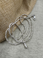 Twisted sterling silver chain necklace coiled against a jute pouch, with the stamped end tag hanging free below the loops.