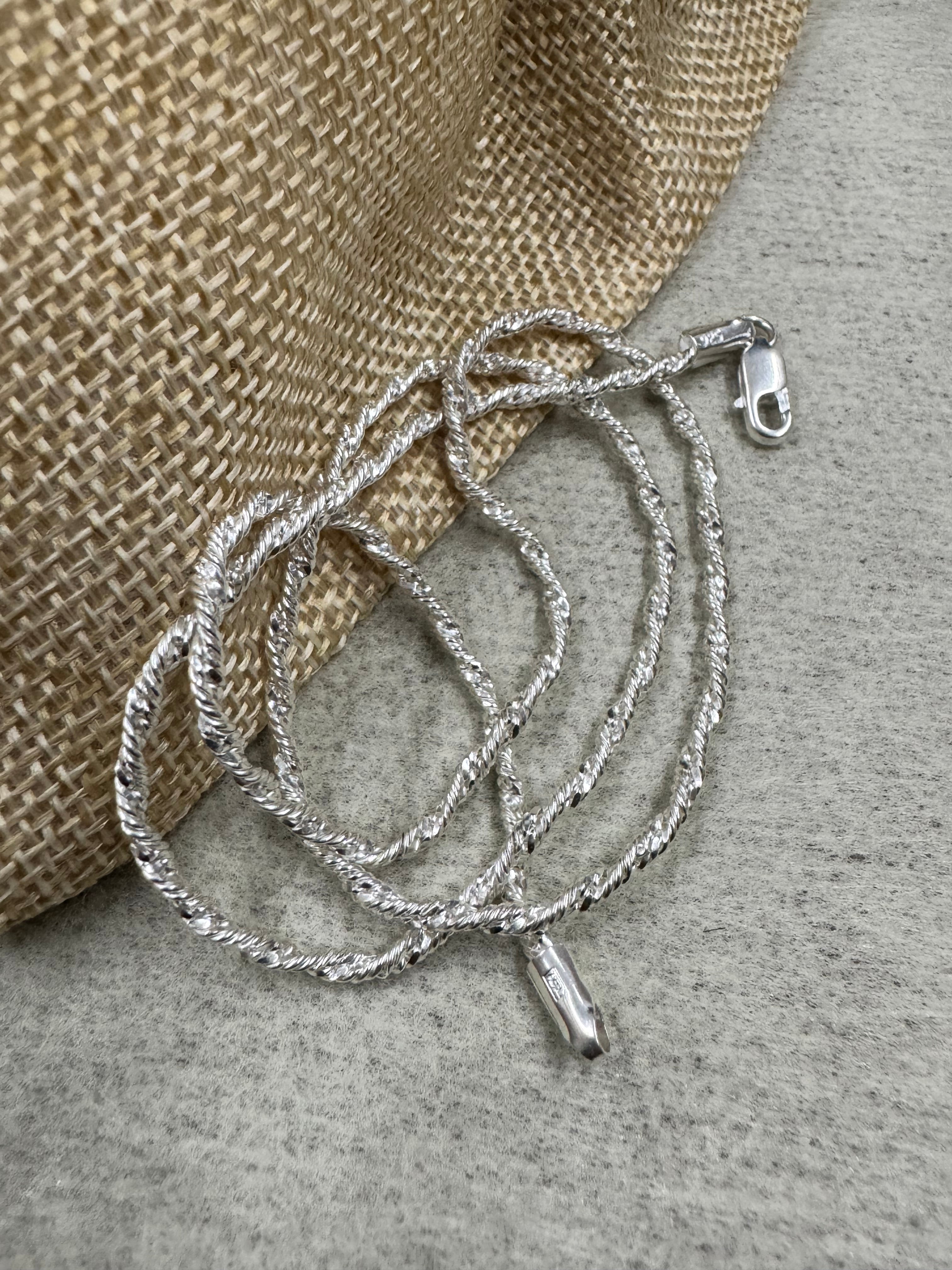 Twisted sterling silver chain necklace coiled against a jute pouch, with the stamped end tag hanging free below the loops.