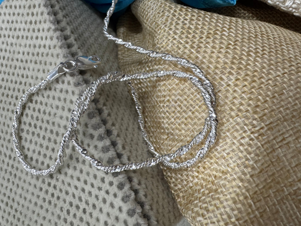 Sterling silver Singapore chain necklace looped over a dotted cream cushion and a jute pouch, lobster clasp at the left.