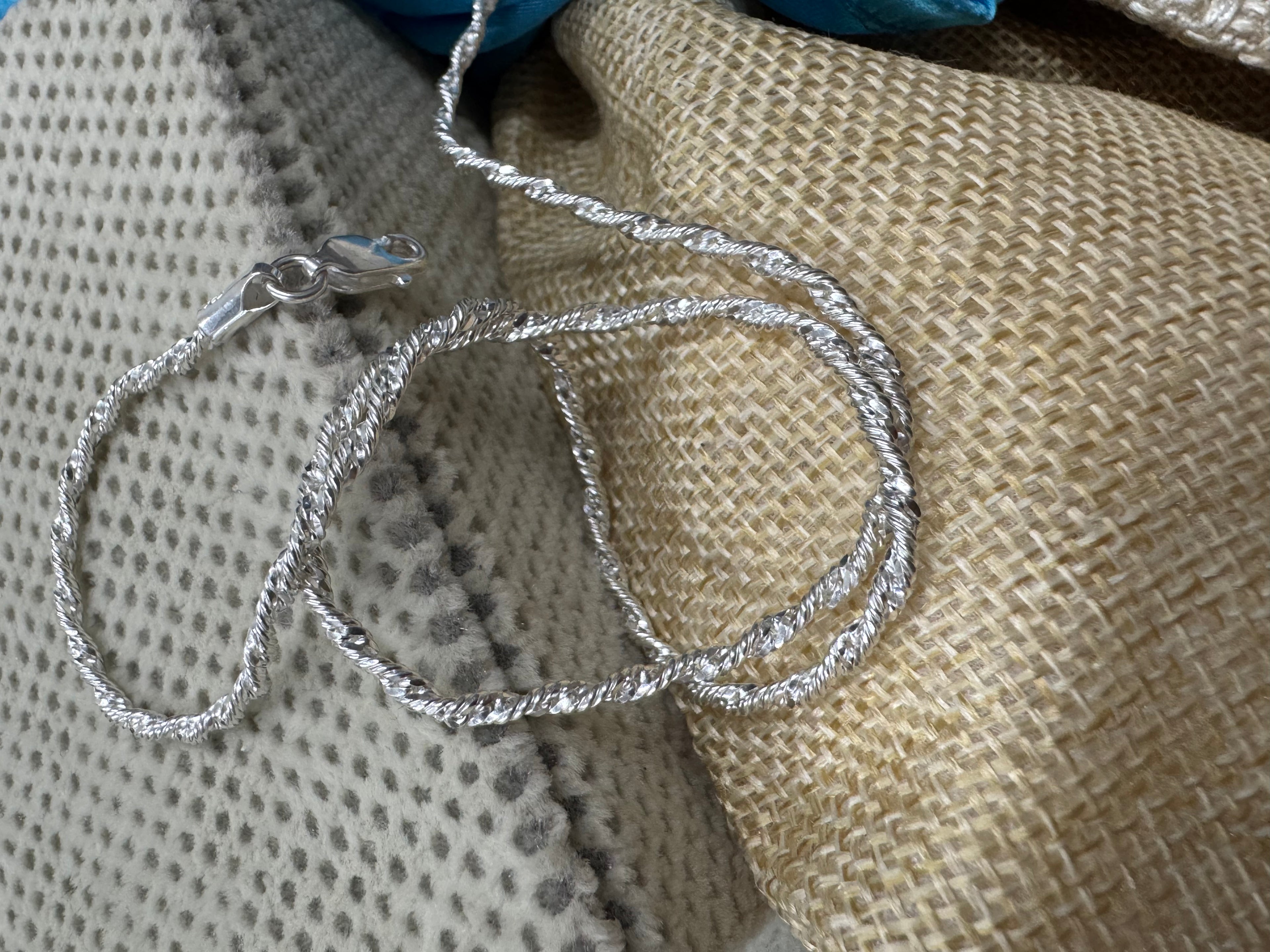 Sterling silver Singapore chain necklace looped over a dotted cream cushion and a jute pouch, lobster clasp at the left.