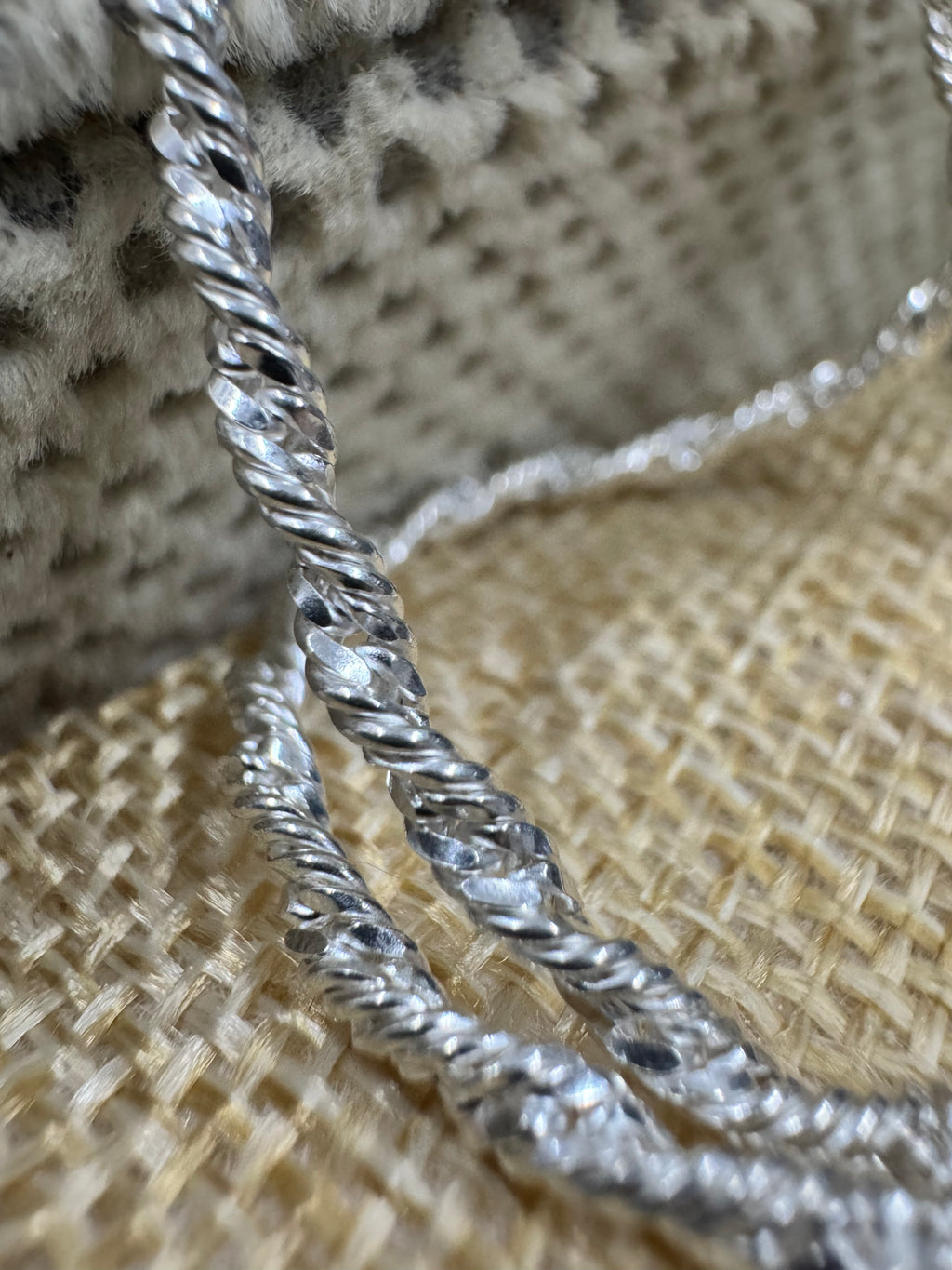Macro view of the twisted, diamond-cut links of the sterling silver Singapore chain against woven jute.