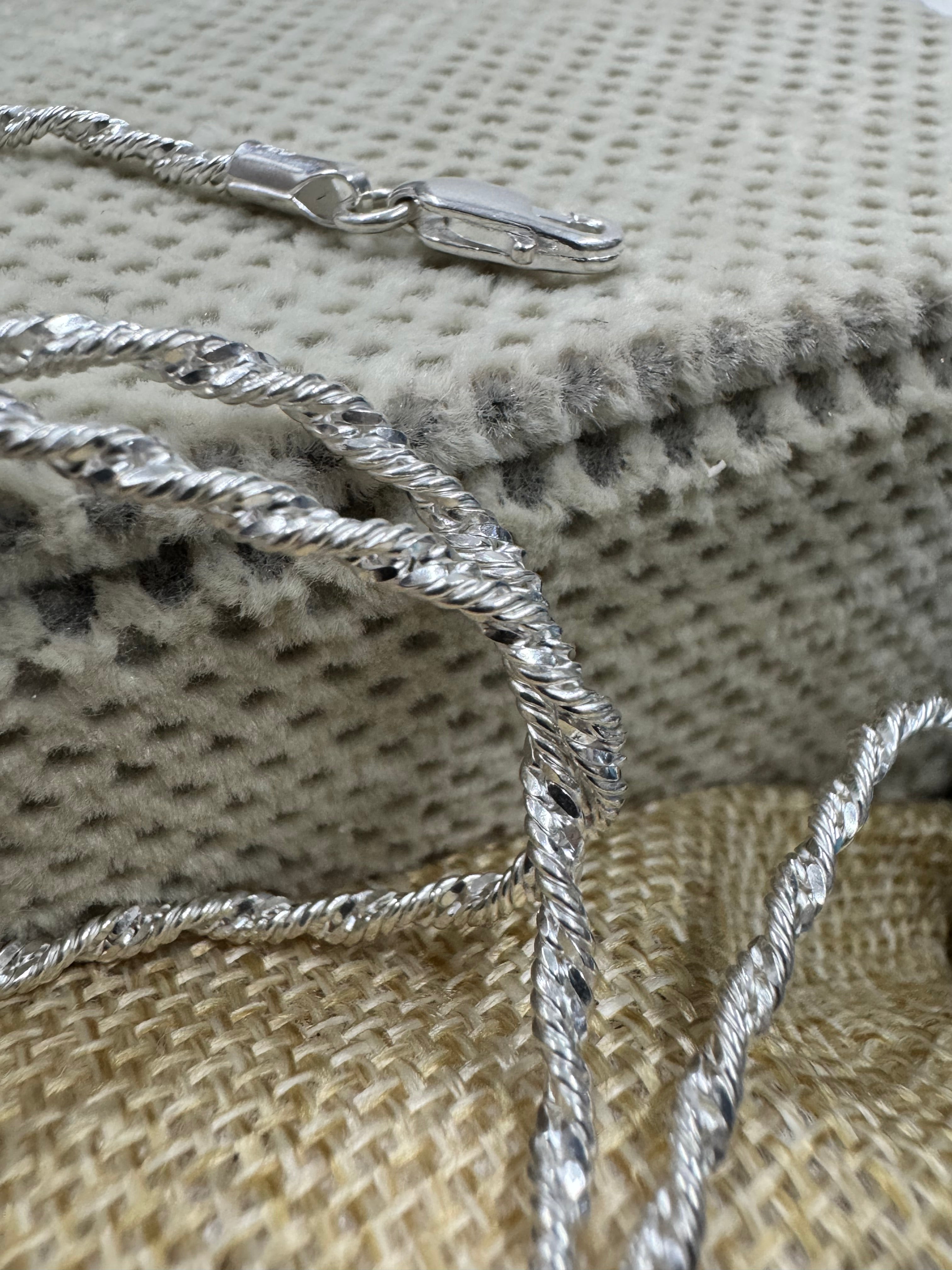 Sterling silver Singapore chain necklace draped over a dotted cream cushion, with the open lobster clasp resting above it.