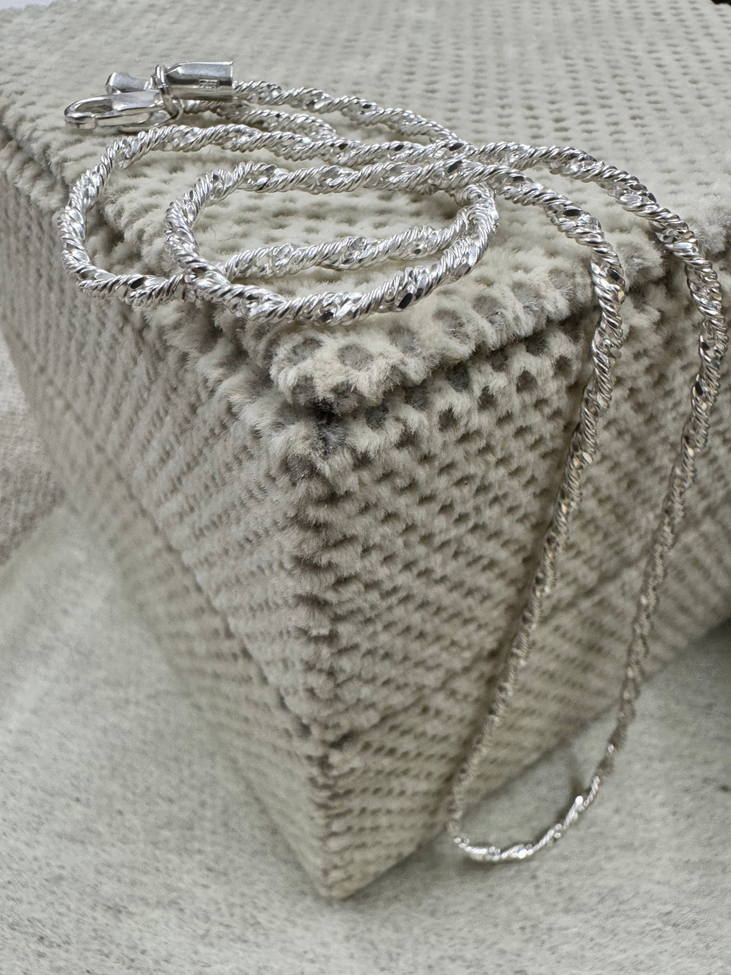 Sterling silver Singapore chain necklace coiled on a cream textured cushion, the clasp at the top and the chain falling over the edge.