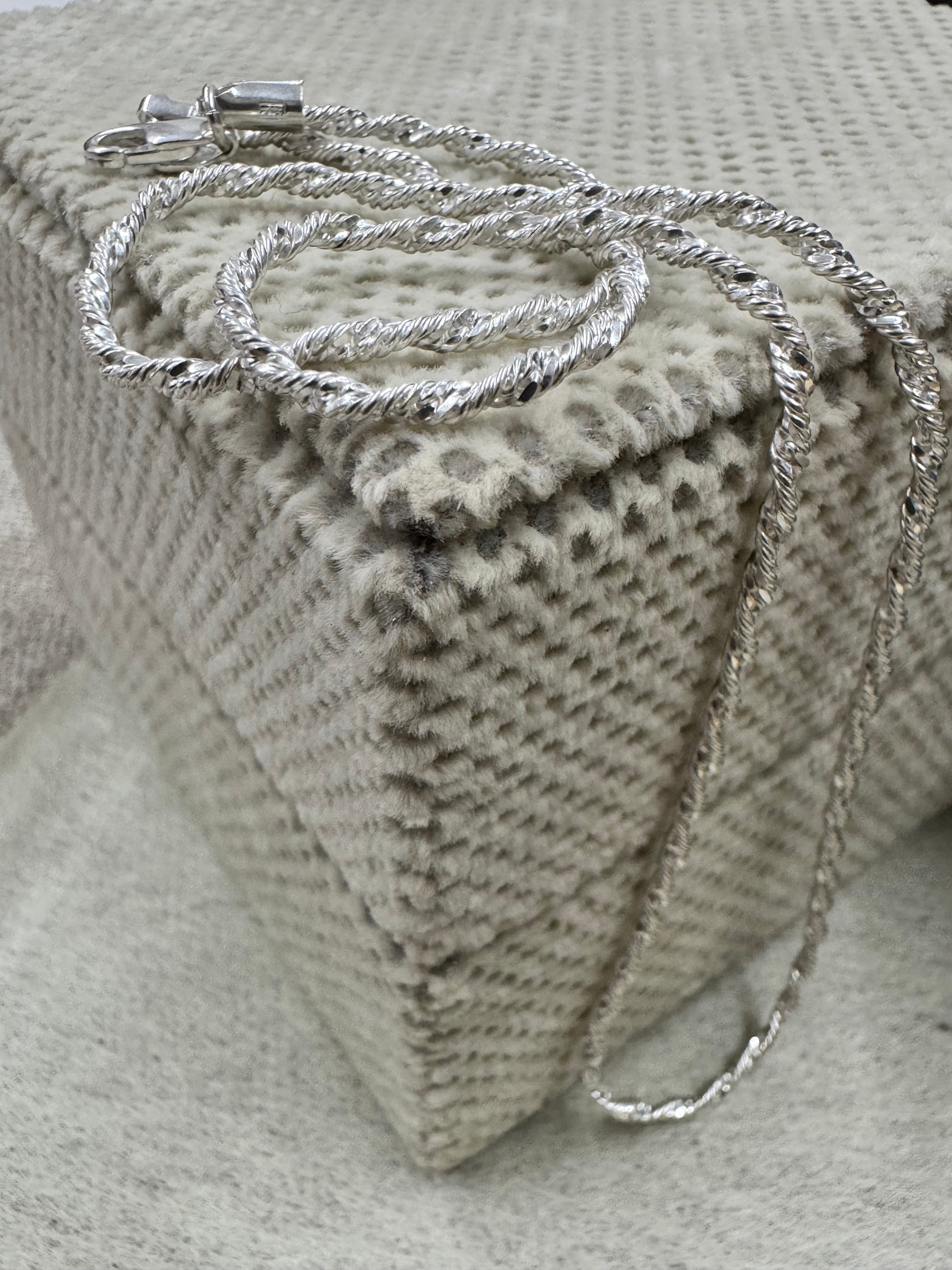 Sterling silver Singapore chain necklace coiled on a cream textured cushion, the clasp at the top and the chain falling over the edge.