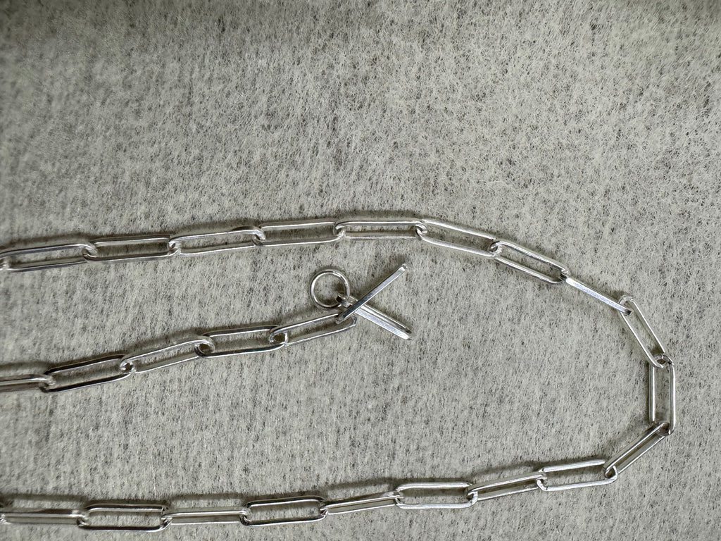 Full length of the sterling silver paperclip chain necklace curved on grey felt, T-bar and ring at the centre.