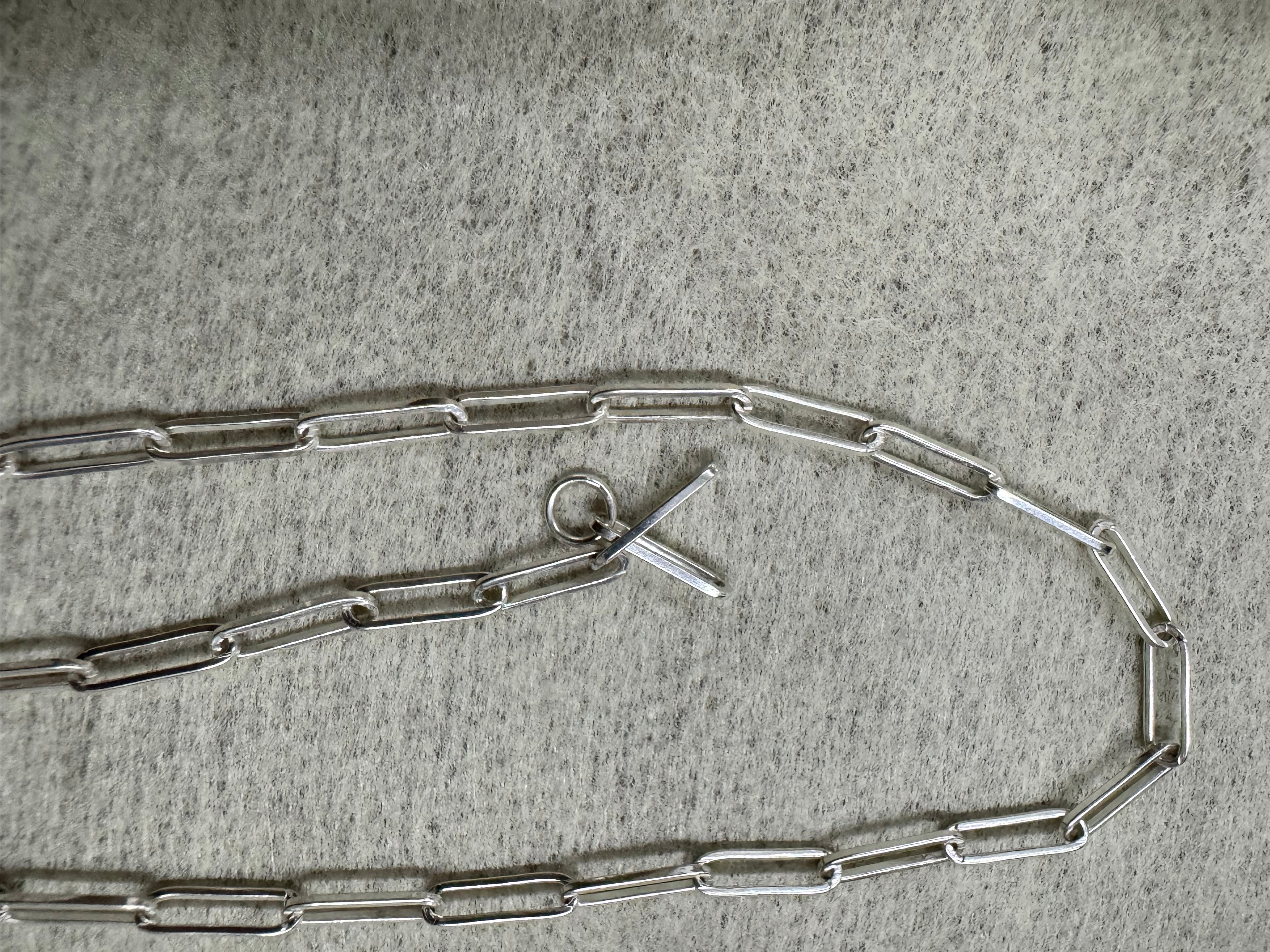 Full length of the sterling silver paperclip chain necklace curved on grey felt, T-bar and ring at the centre.