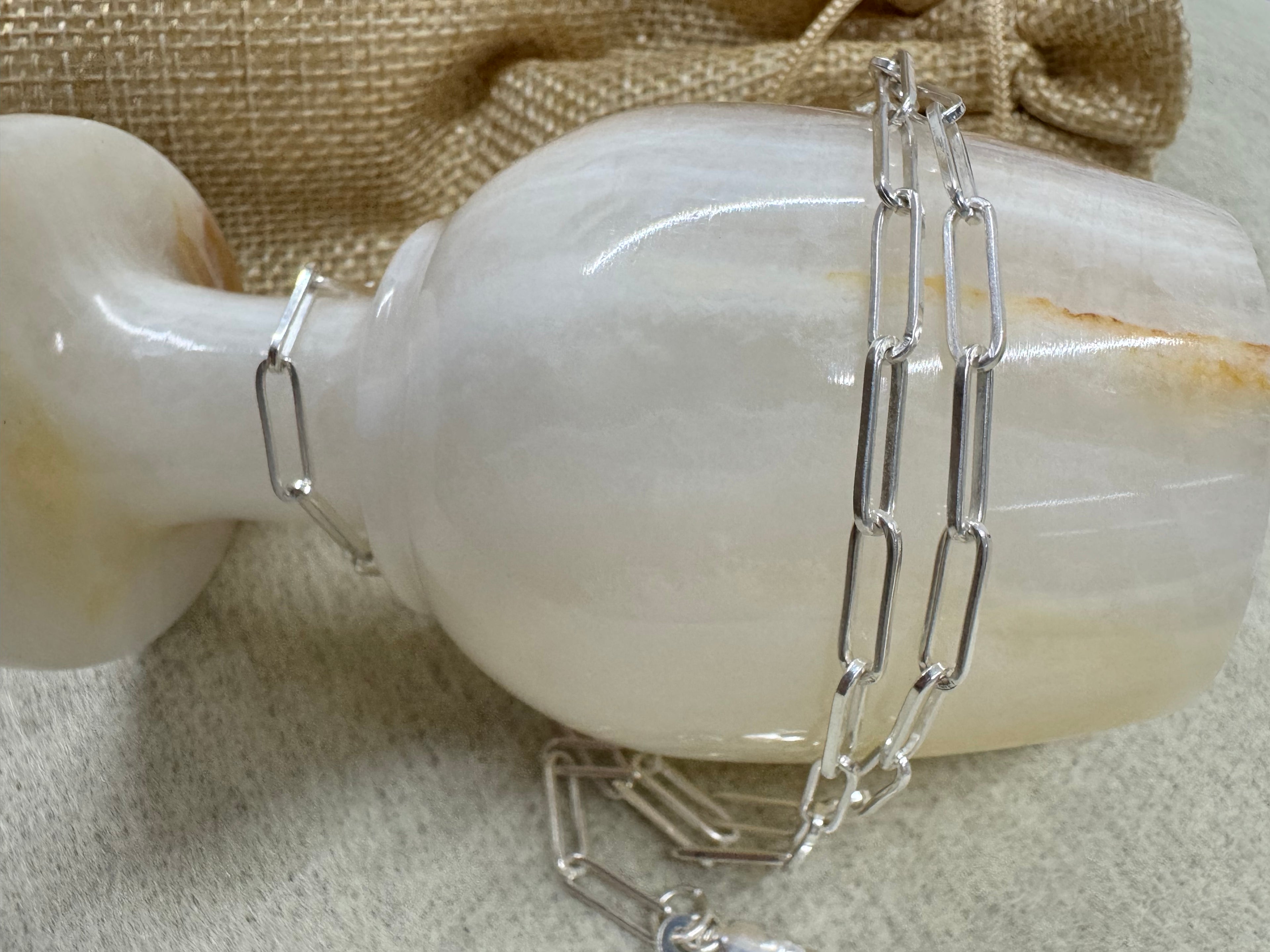 Sterling silver paperclip chain necklace draped over a pale polished vase beside a jute pouch.