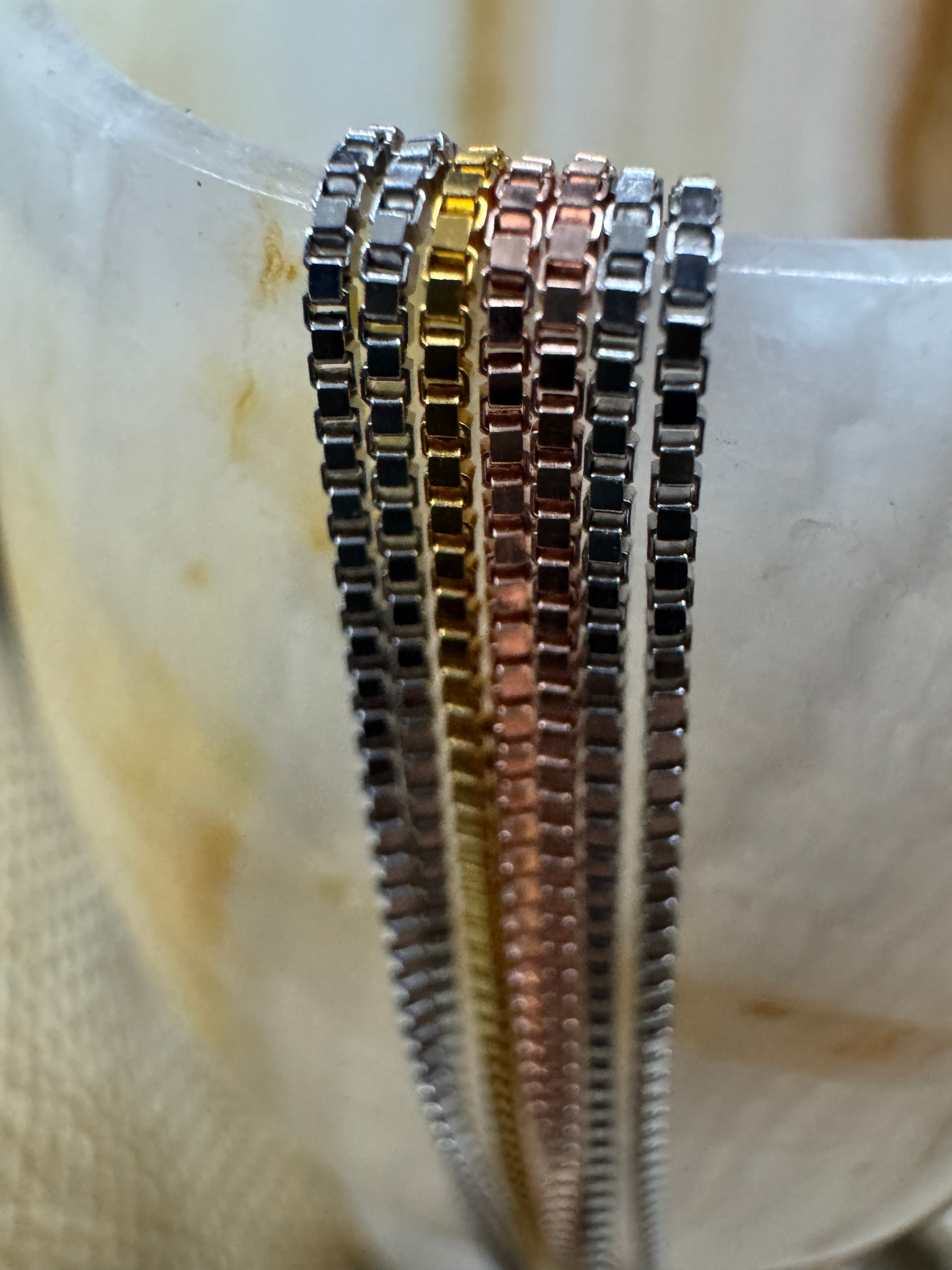 Macro view of the box links of the sterling silver necklace laid over a pale polished vase, showing silver, gold-tone, rose-tone and oxidised strands.