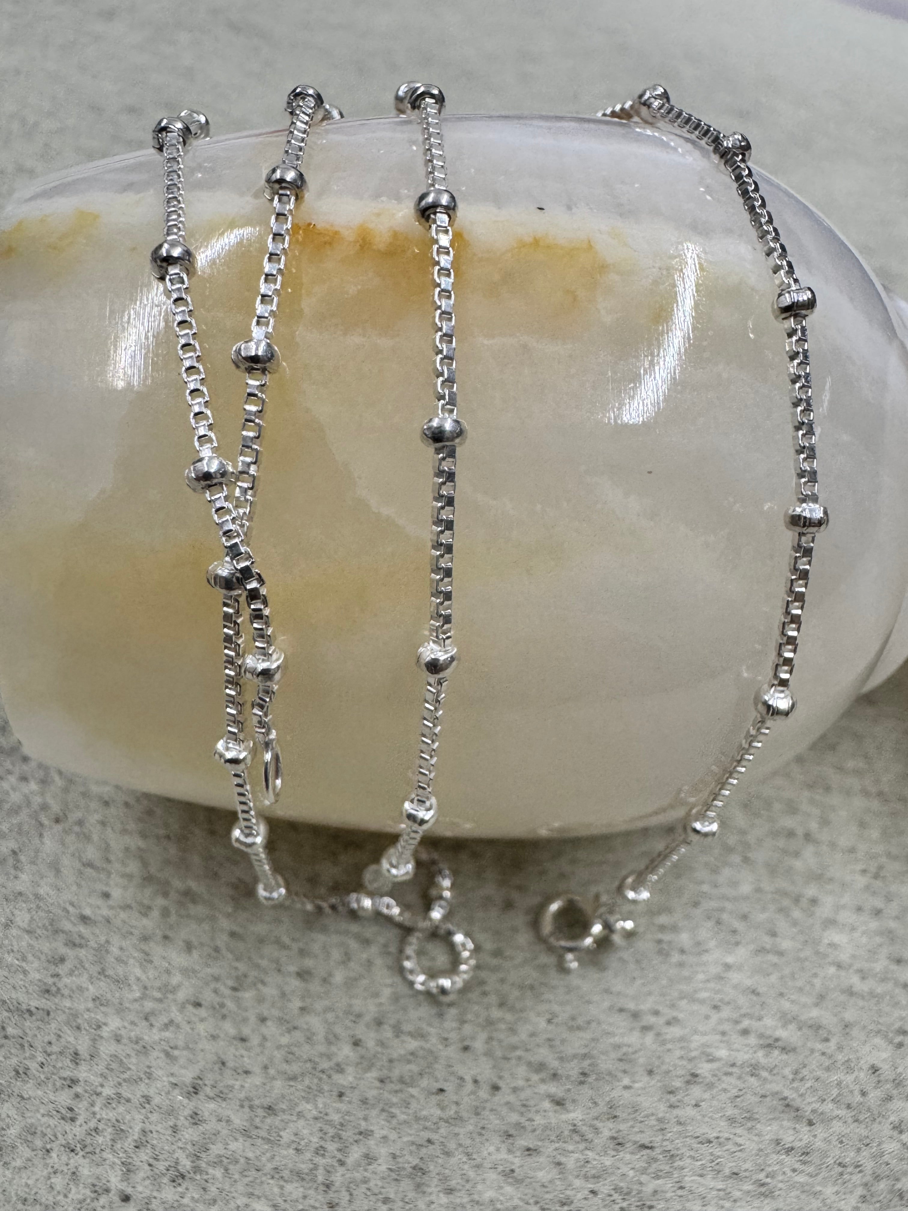 The sterling silver box chain necklace draped over a pale banded stone, showing the beads spaced along the line.