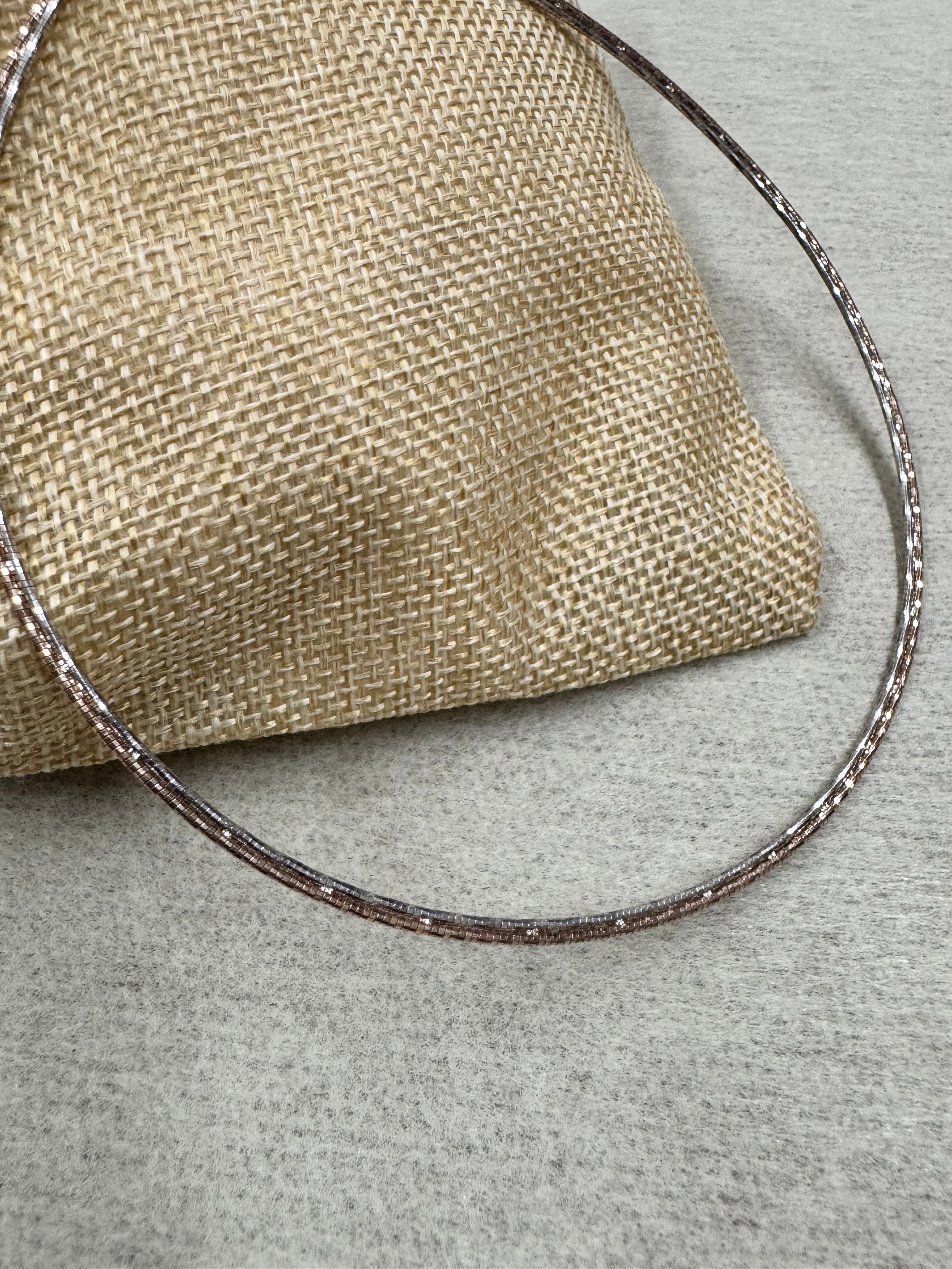 The sterling silver snake chain necklace curving over a jute pouch, its diamond-cut facets catching light.