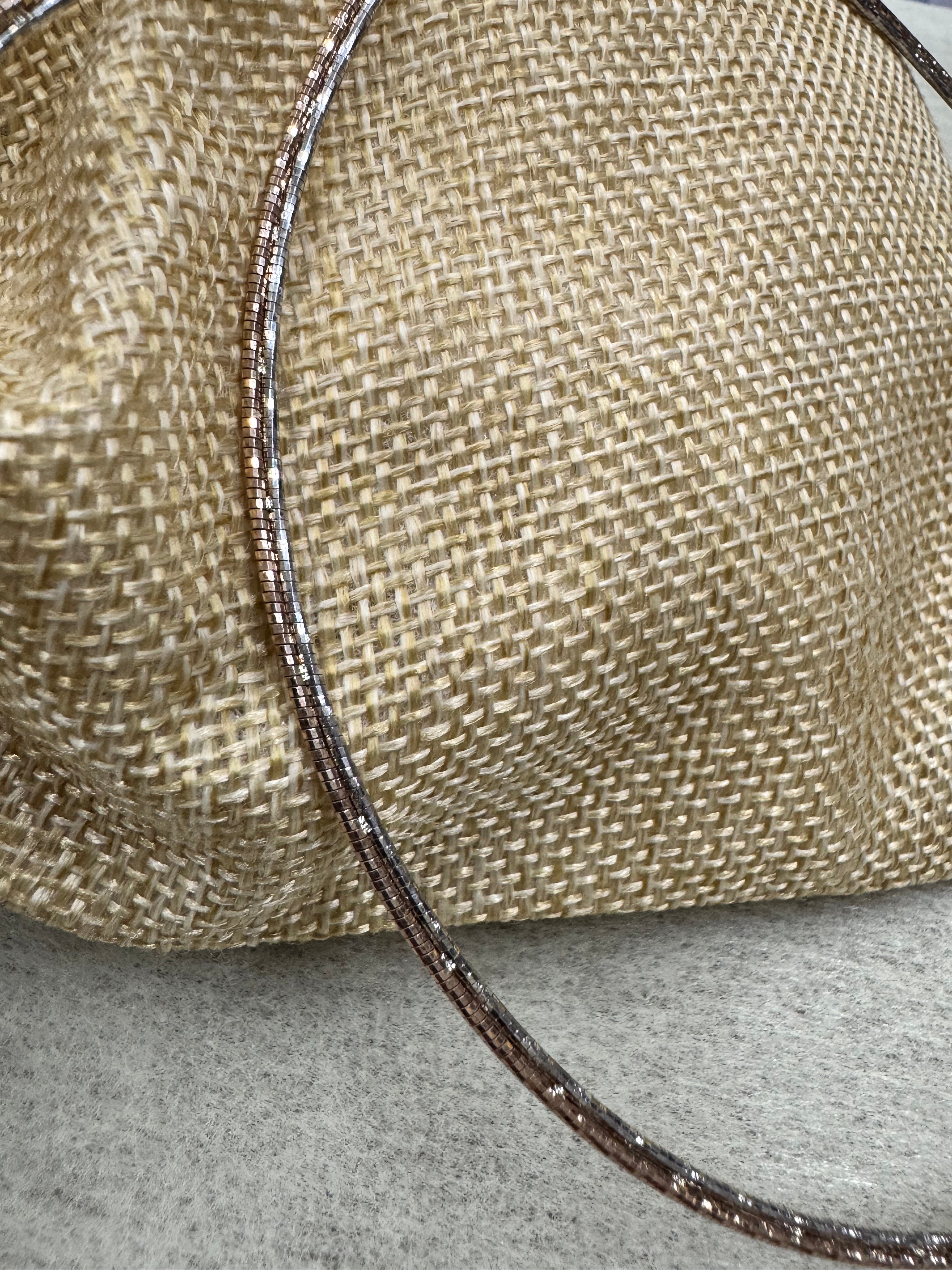 The sterling silver snake chain necklace arcing across woven jute as one continuous diamond-cut line.