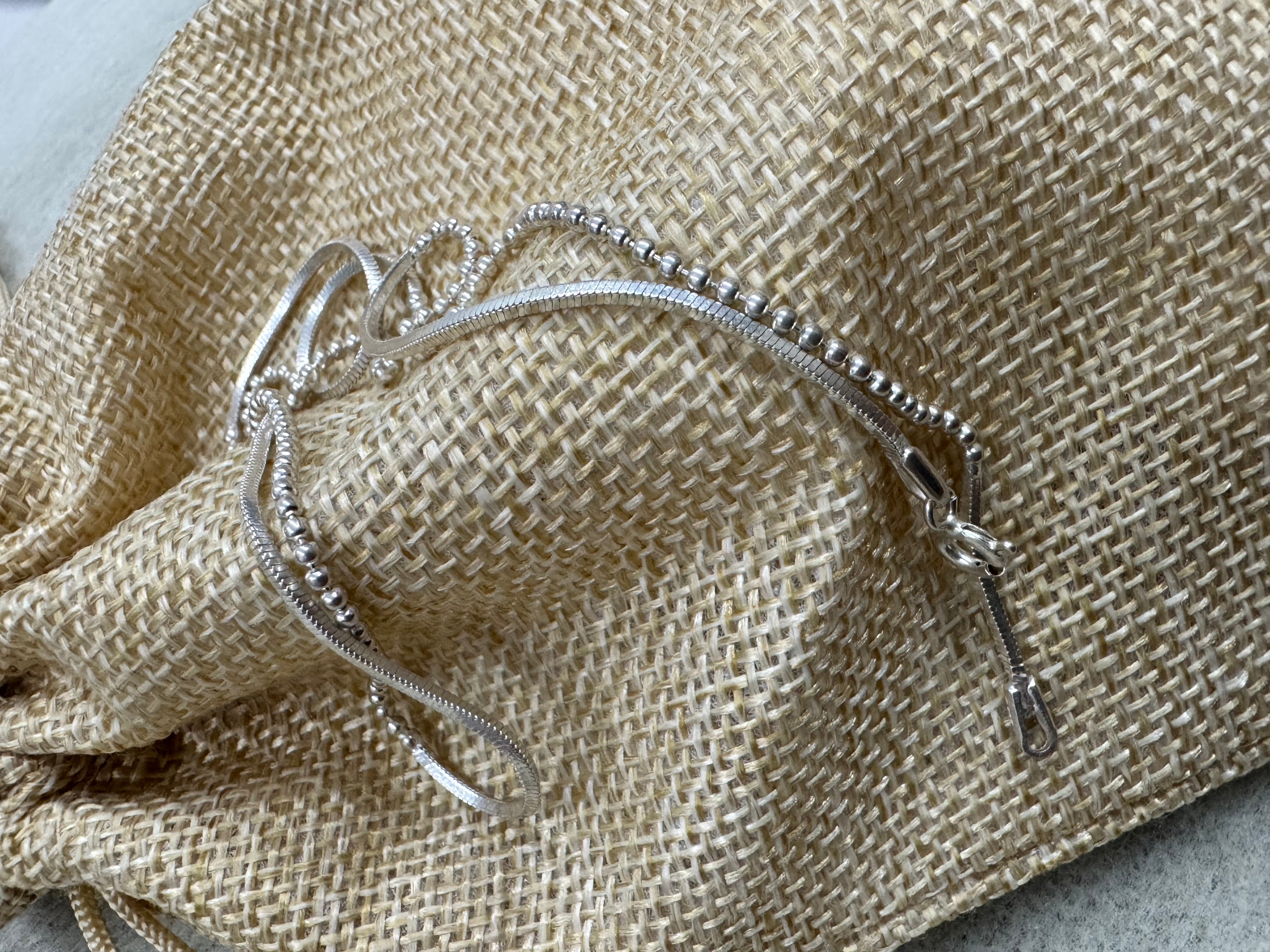 Sterling silver chain necklace resting on a woven jute pouch, showing a bead section running into a snake section beside the flat end tag.