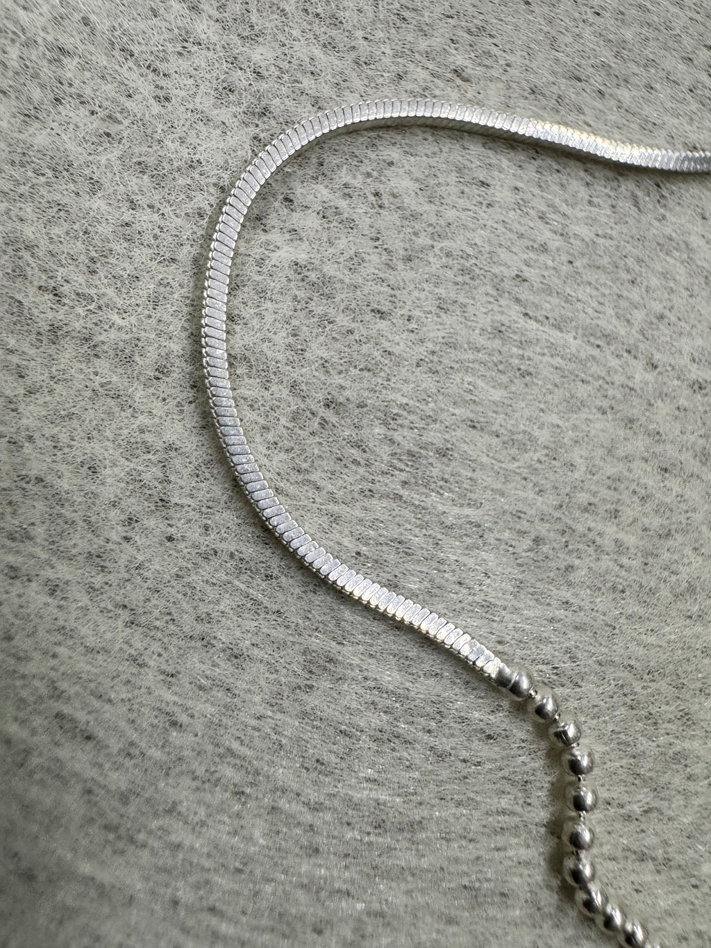 Close view of a snake section of the sterling silver chain necklace running into a row of round beads on grey felt.