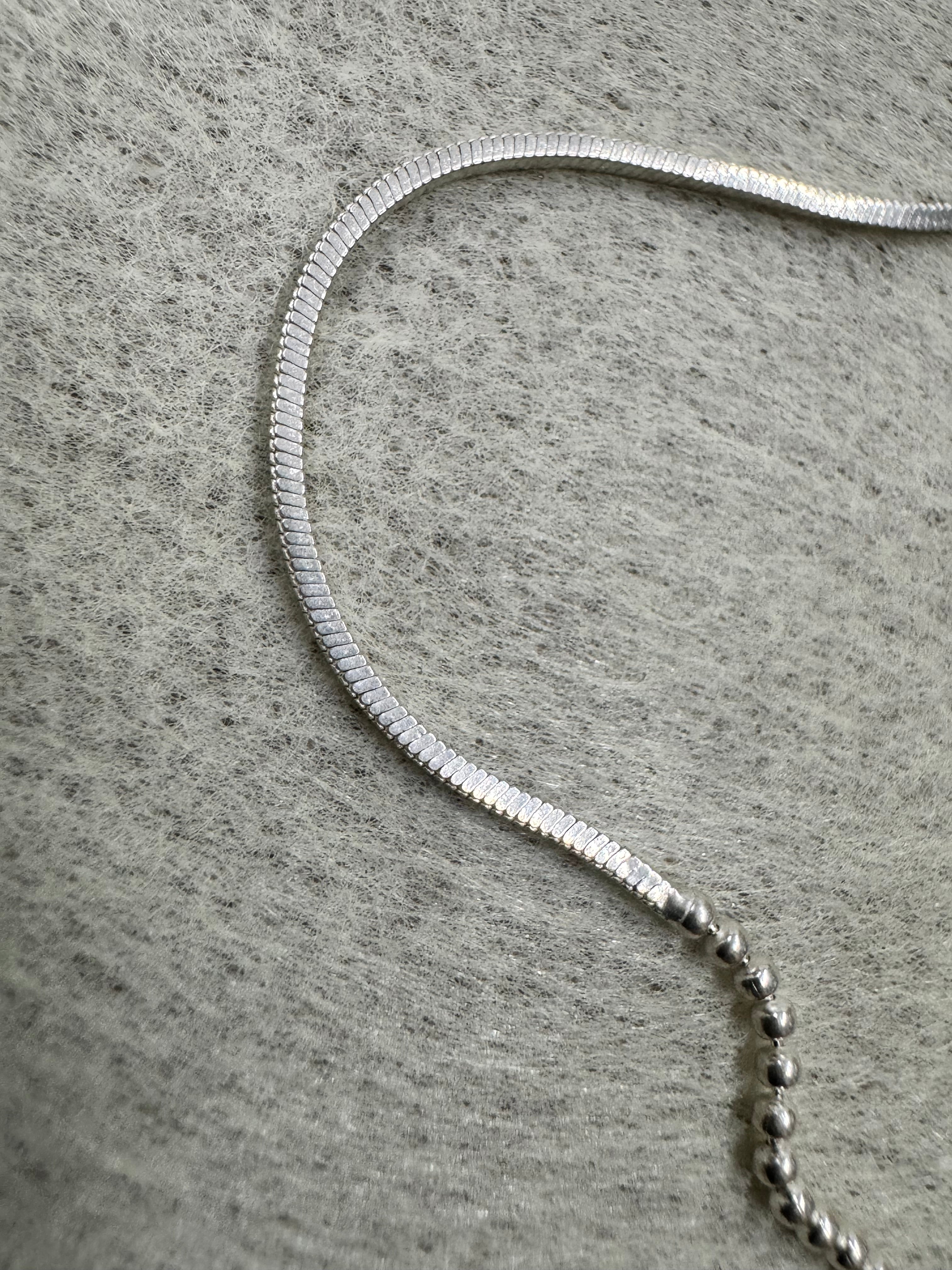 Close view of a snake section of the sterling silver chain necklace running into a row of round beads on grey felt.