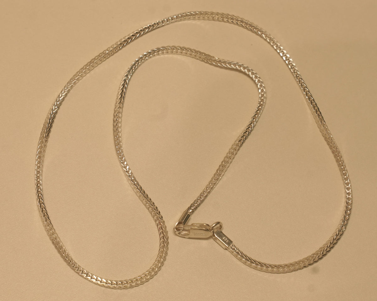 Slim sterling silver foxtail chain coiled on a pale surface with the clasp at the centre.