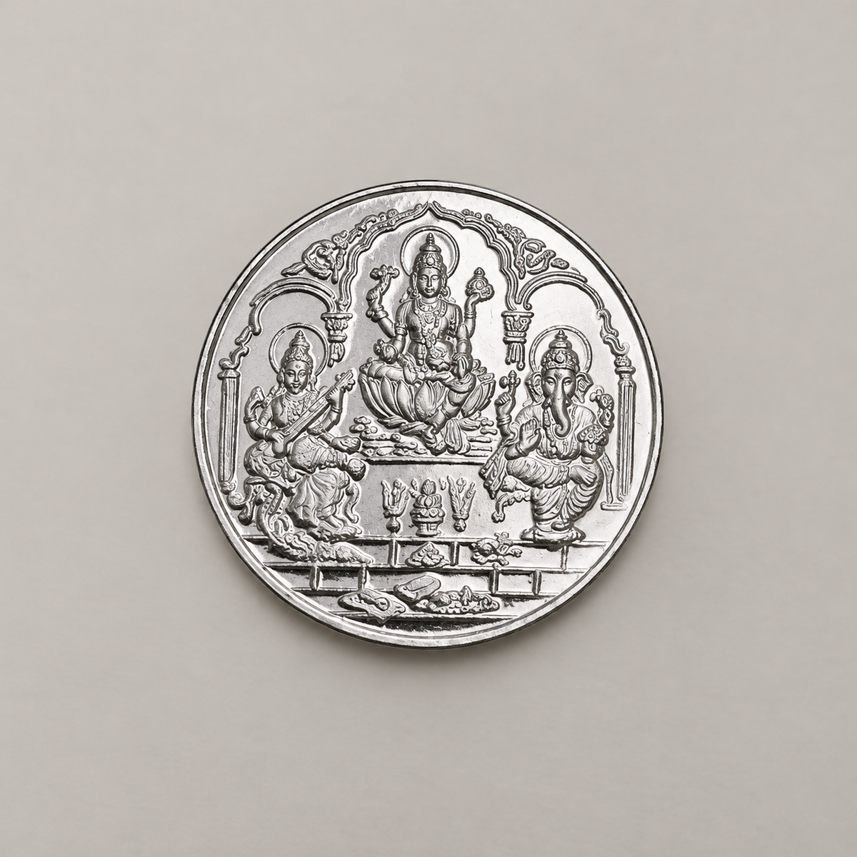 20 gram fine silver coin with Lakshmi, Ganesh and Saraswati under temple arches, on a beige surface.