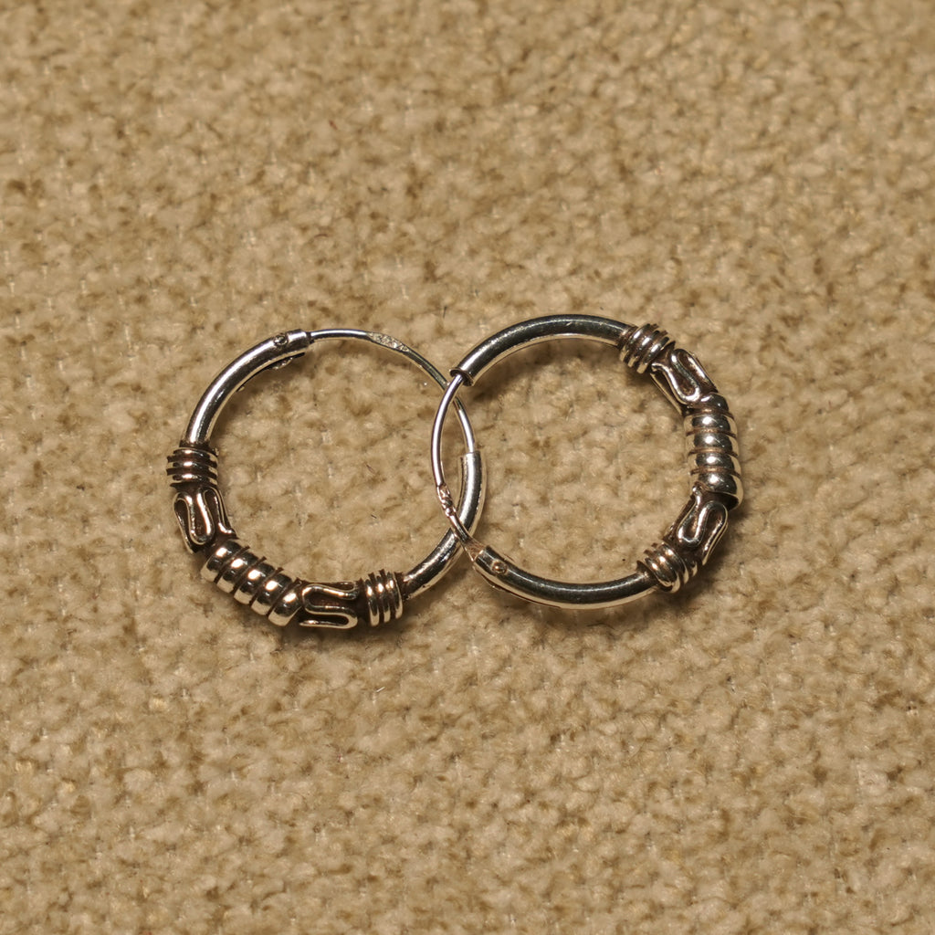 Pair of sterling silver Bali hoop earrings with coiled bands and scroll motifs, overlapping on beige fabric.