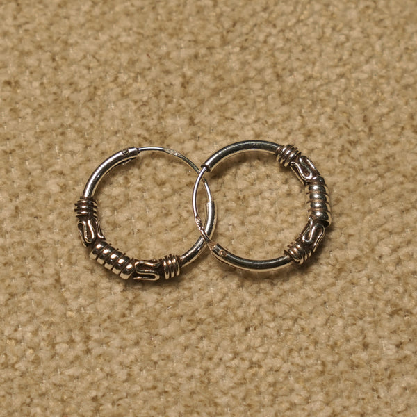 Pair of sterling silver Bali hoop earrings with coiled bands and scroll motifs, overlapping on beige fabric.