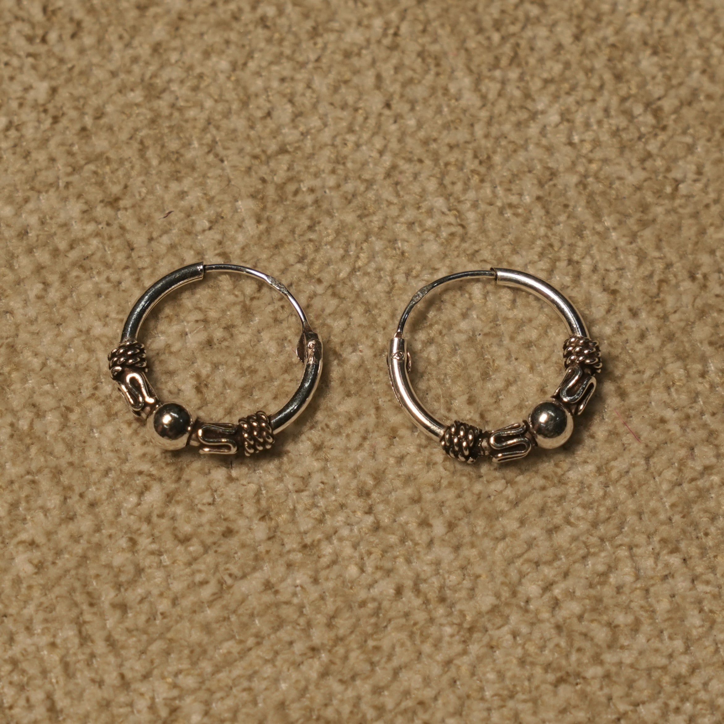 Pair of sterling silver Bali hoop earrings with a central polished bead flanked by rope coils, on beige fabric.