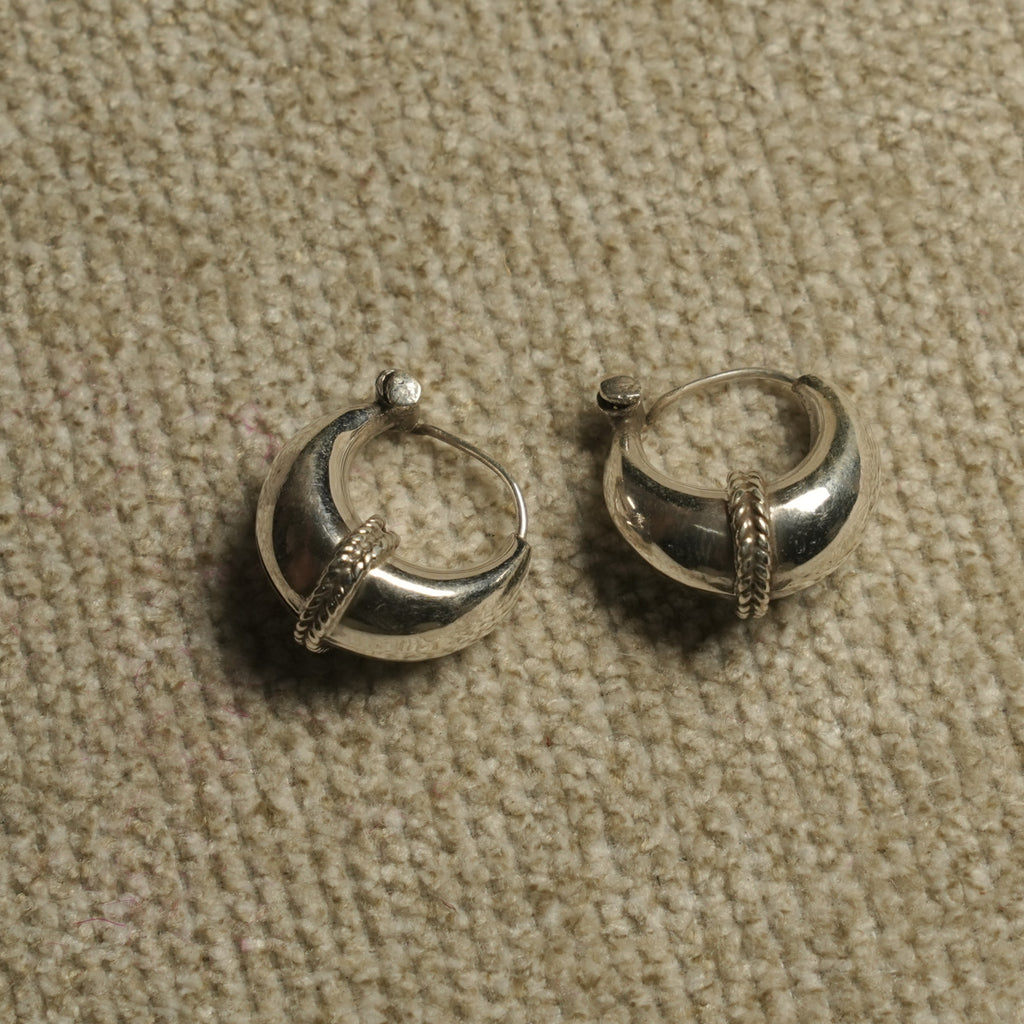 Pair of polished sterling silver dome hoop earrings with a rope-textured band across the front, resting on beige woven fabric.