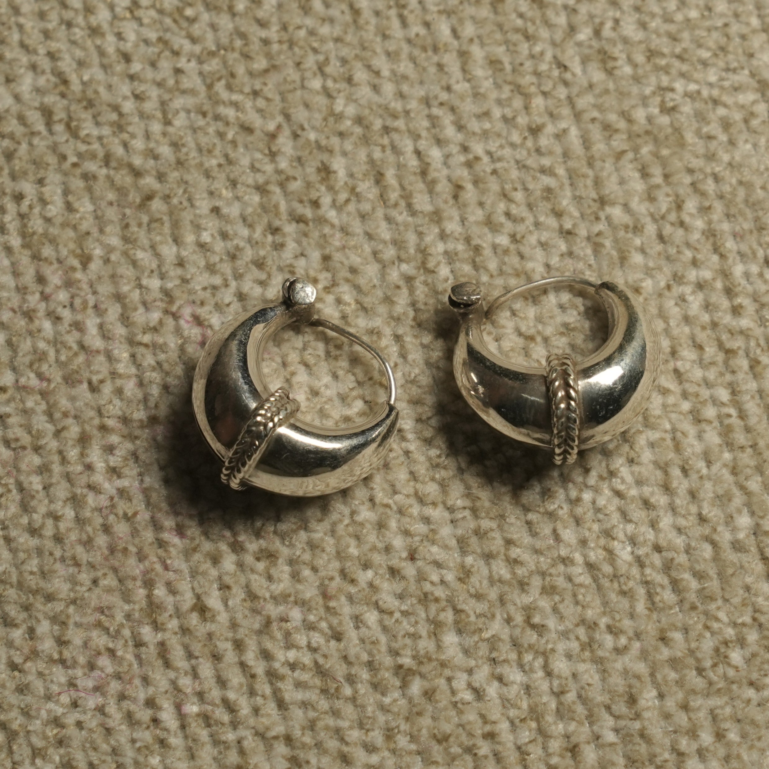 Pair of polished sterling silver dome hoop earrings with a rope-textured band across the front, resting on beige woven fabric.