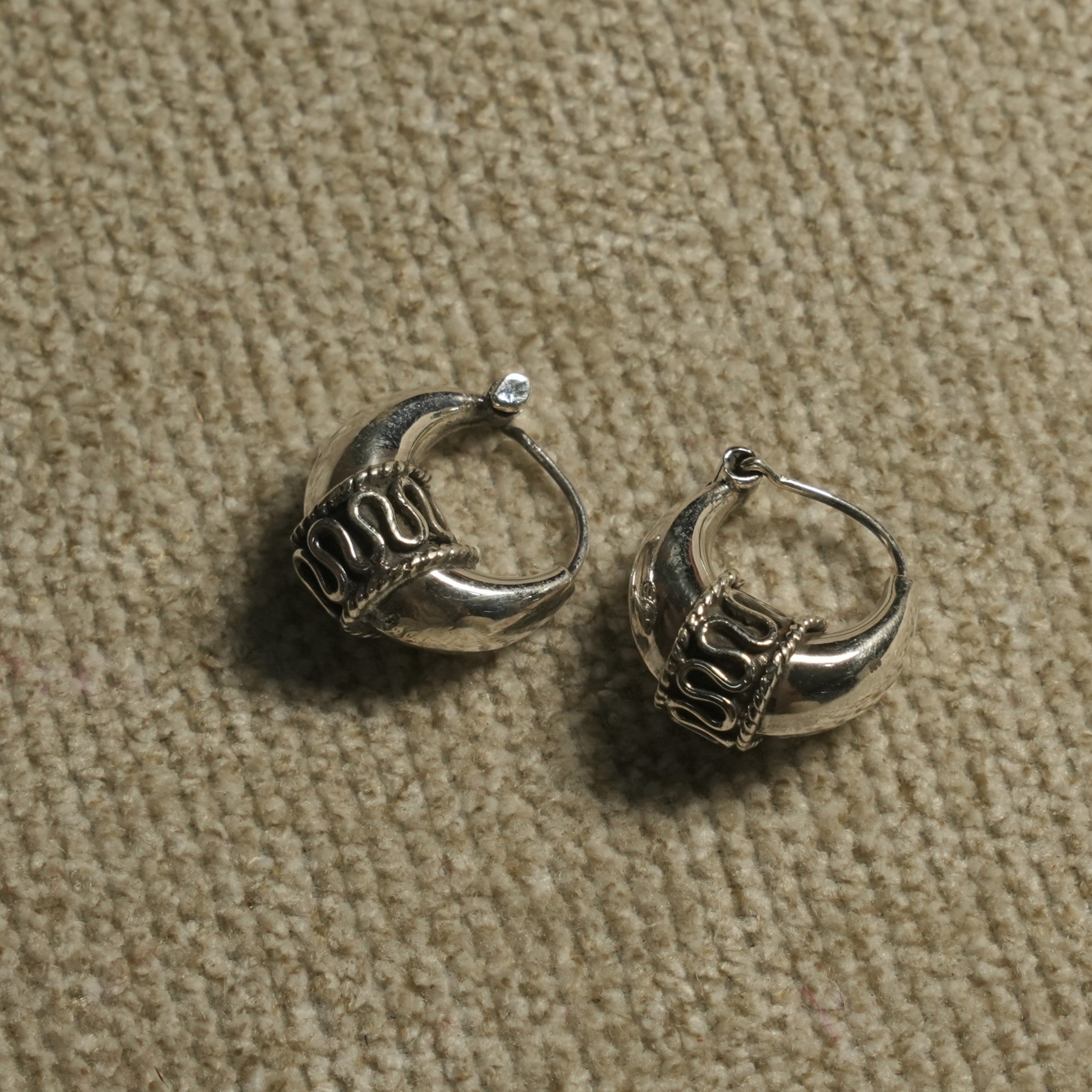 Close view of the sterling silver dome hoops showing the oxidised scrollwork band with rope edging and the hinged wire closure, on beige woven fabric.