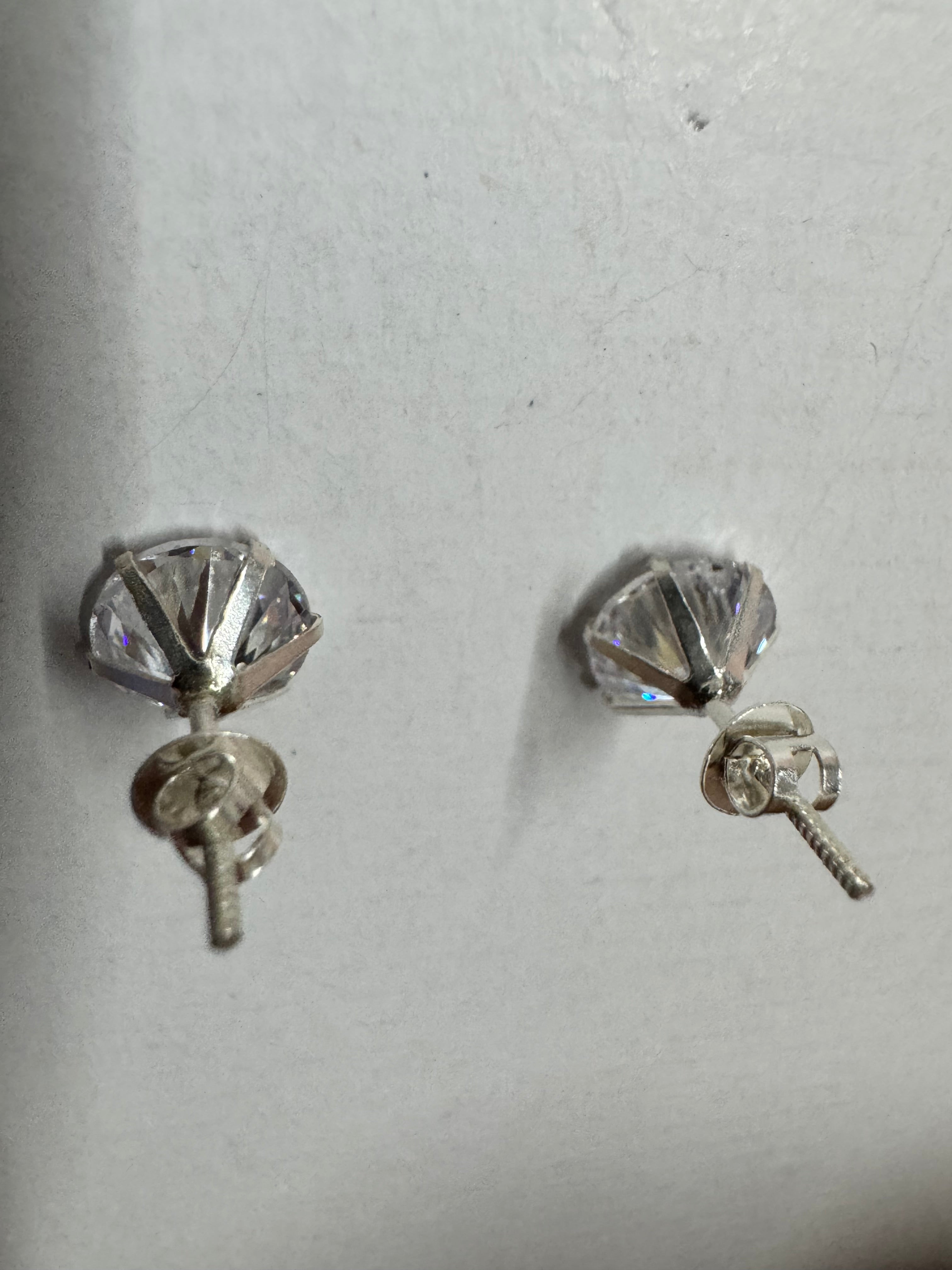 Underside of the two sterling silver stud earrings on a pale grey surface, the open claw baskets showing the pavilion of each cubic zirconia with the screw-on backs run part-way down the threaded posts.
