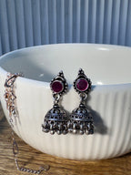 Pair of oxidised sterling silver jhumkas with deep pink glass stone tops, resting on a plain background.