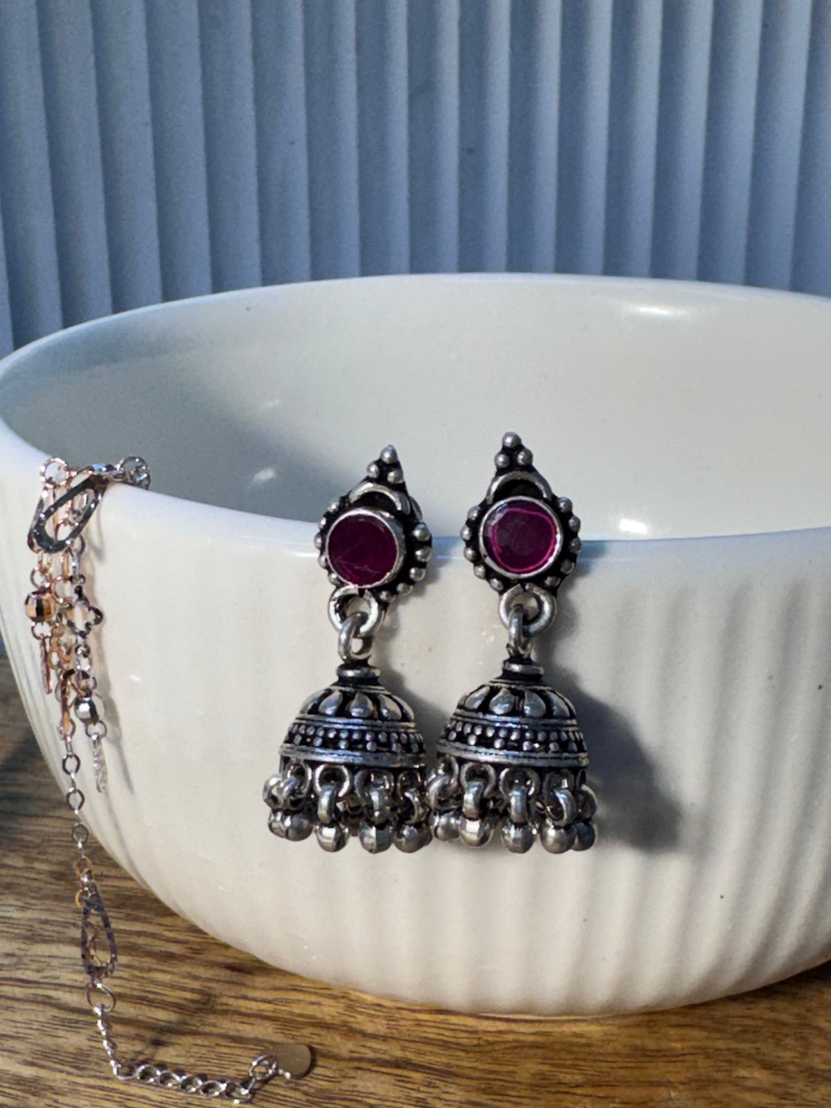 Pair of oxidised sterling silver jhumkas with deep pink glass stone tops, resting on a plain background.