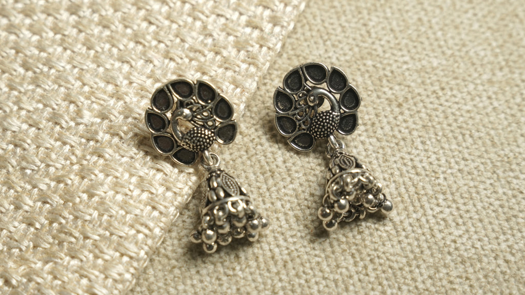 Pair of oxidised sterling silver peacock jhumka earrings with teardrop cutwork tops and beaded bell drops, on ivory woven fabric.