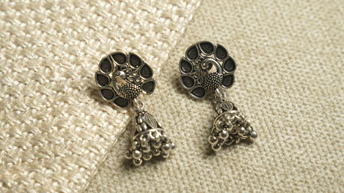 Pair of oxidised sterling silver peacock jhumka earrings with teardrop cutwork tops and beaded bell drops, on ivory woven fabric.